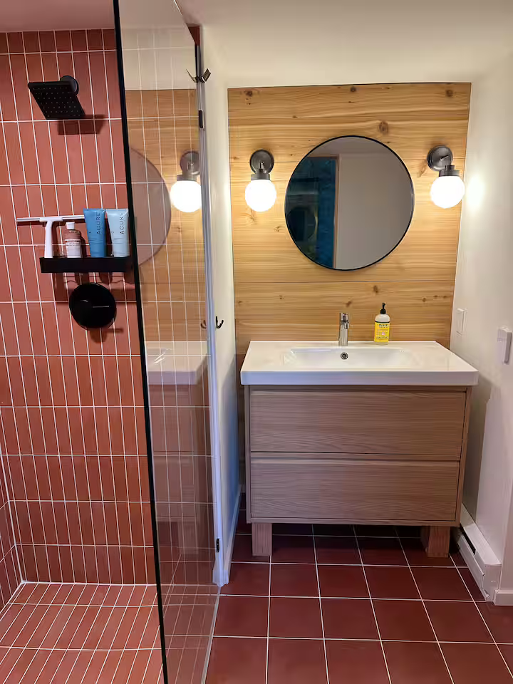 Bathroom with a walk-in shower area with red tiles, a wooden vanity with a sink, a round mirror on a wooden wall, and wall-mounted lights.