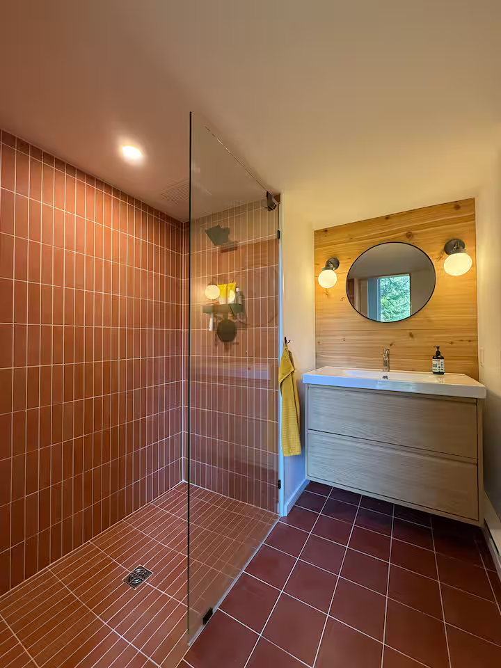 A modern bathroom with a walk-in shower with red tile walls and floor, a wood-paneled wall with a mirror, and a vanity with a sink, lit by wall sconces.