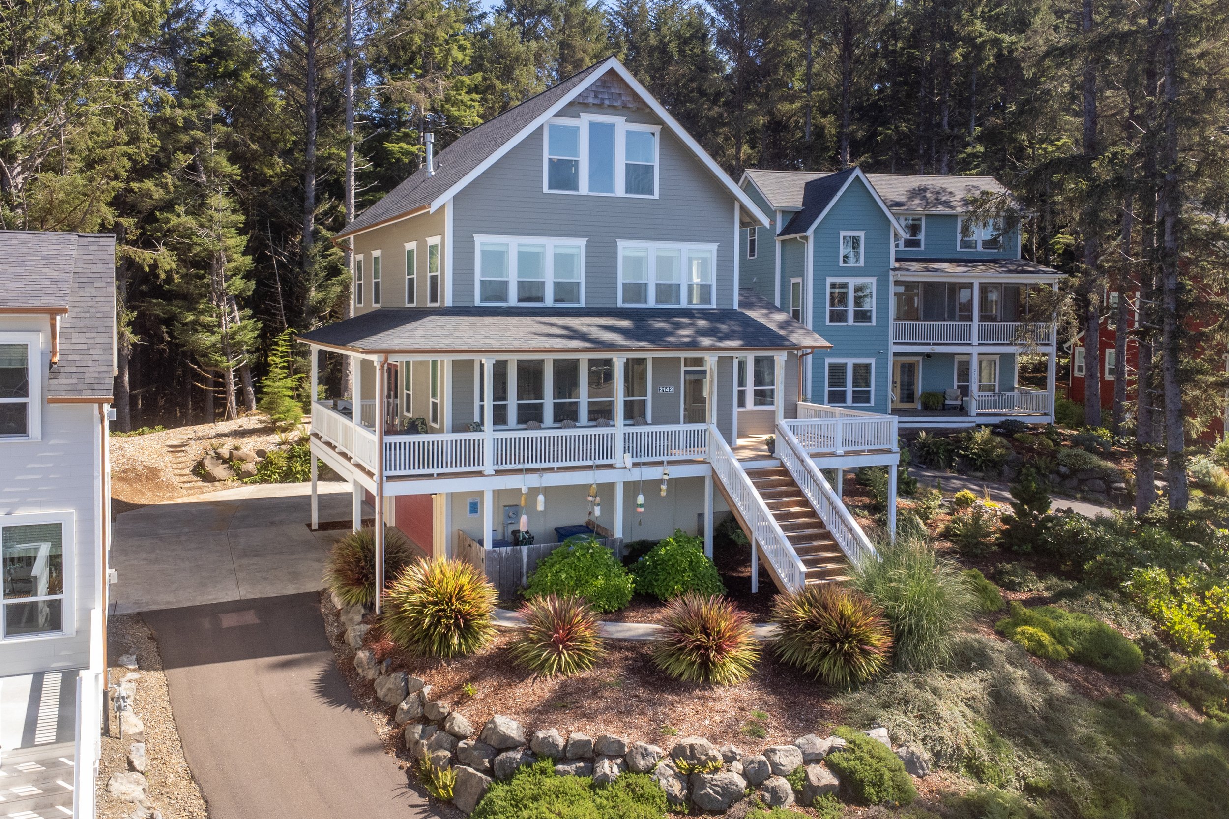 A large, modern house painted in light blue with multiple large windows, situated in a wooded area with tall pine trees surrounding it. The house has a front porch with white railings and a staircase leading up to it. There is a paved pathway in front, bordered by rocks and landscaped plants, and the sky is clear and sunny.
