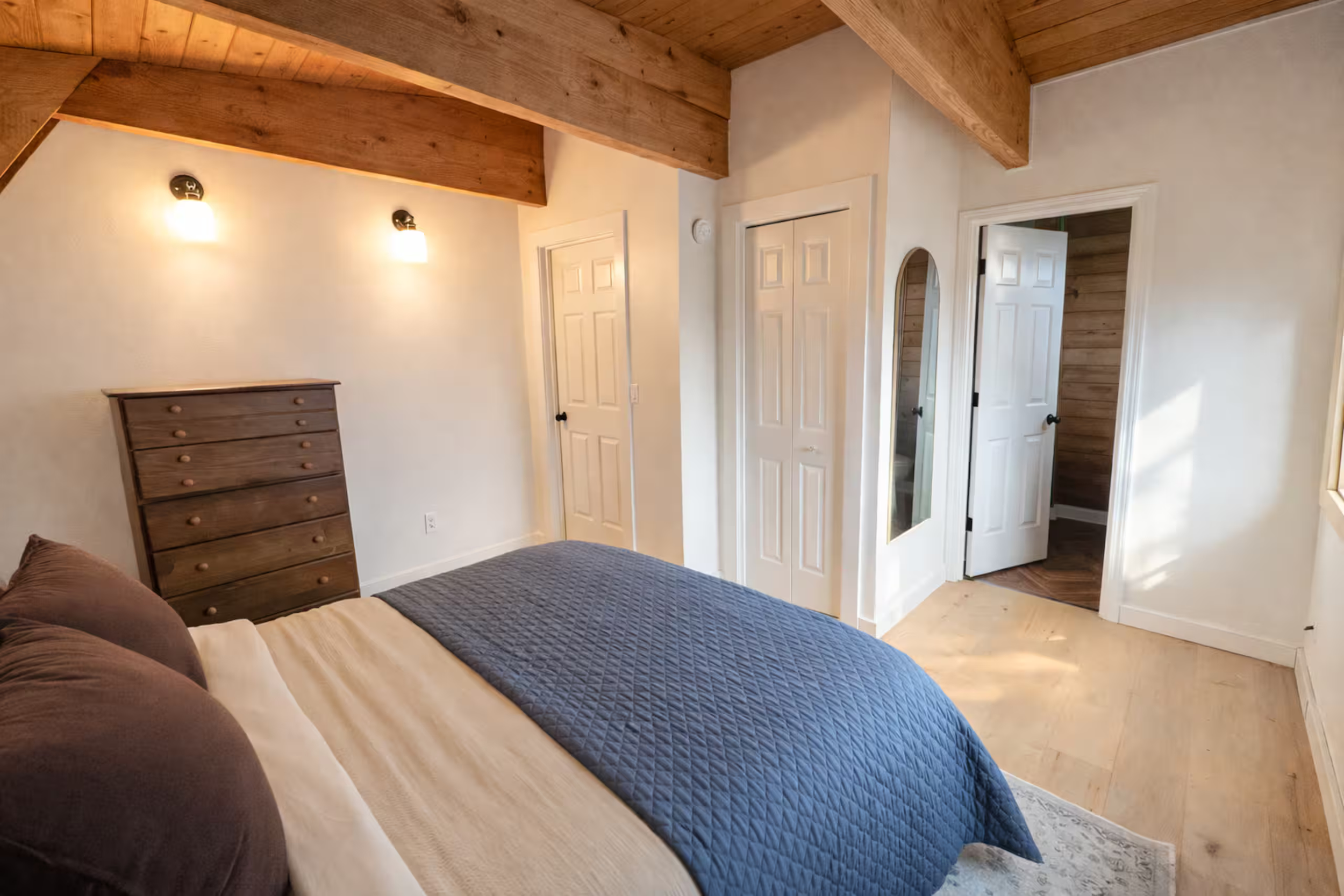 A cozy bedroom with a bed, wooden dresser, and bathroom doors, featuring wooden ceiling beams and light-colored walls.
