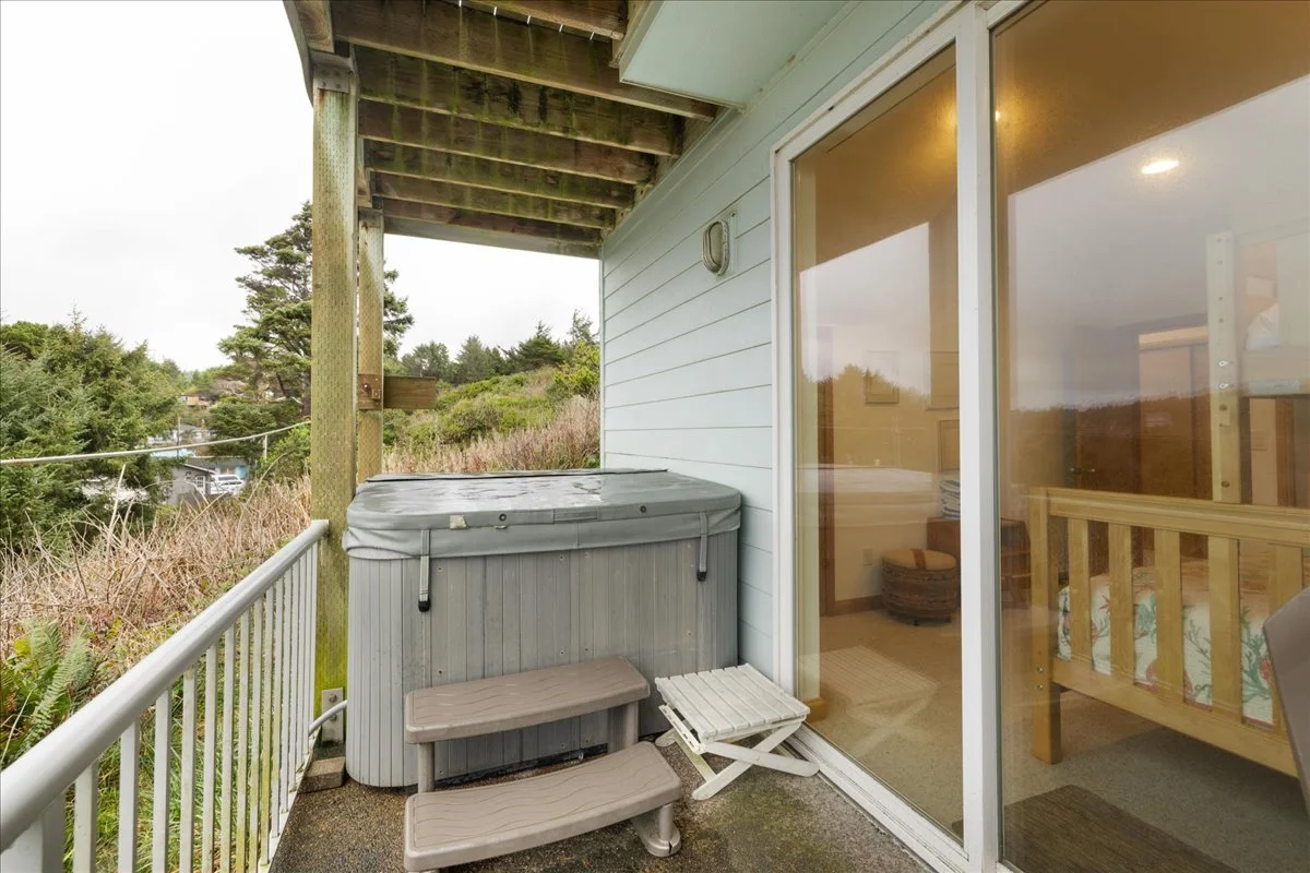 Balcony with hot tub, two beige steps, sliding glass door, wooden railing, and view of trees and neighborhood.
