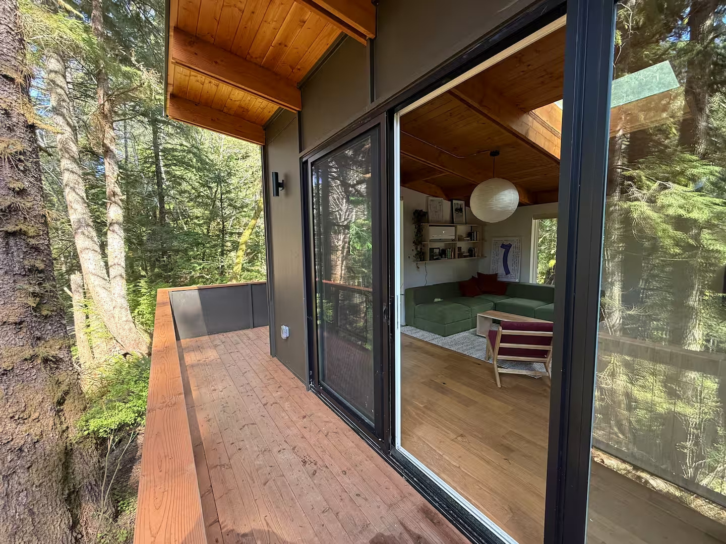View of a modern house with a wooden balcony and glass sliding doors, overlooking a wooded forest area.