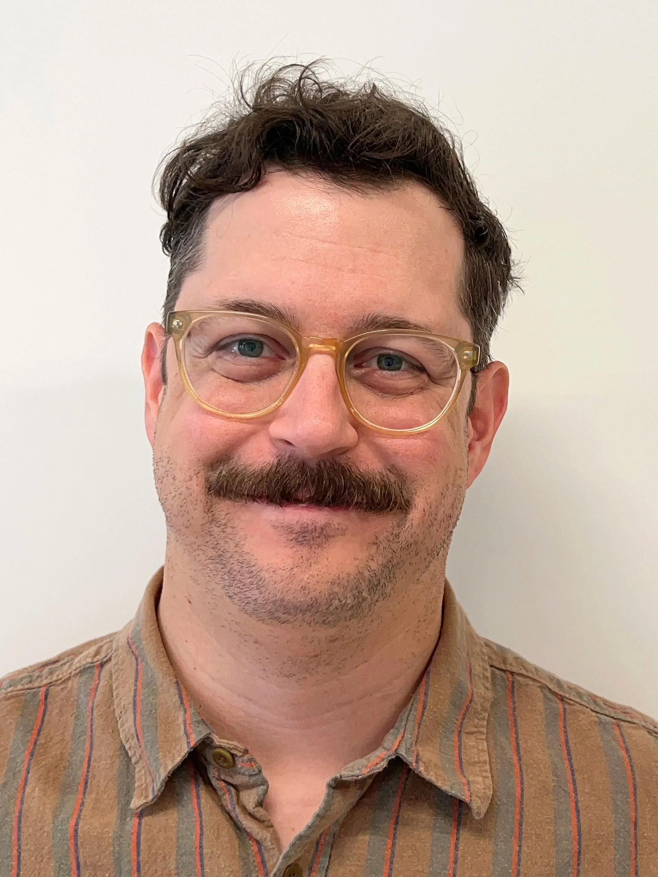 Close-up photo of a man with dark, curly hair, green eyes, glasses, a mustache, and a beard, wearing a brown striped button-up shirt, standing against a plain white wall.