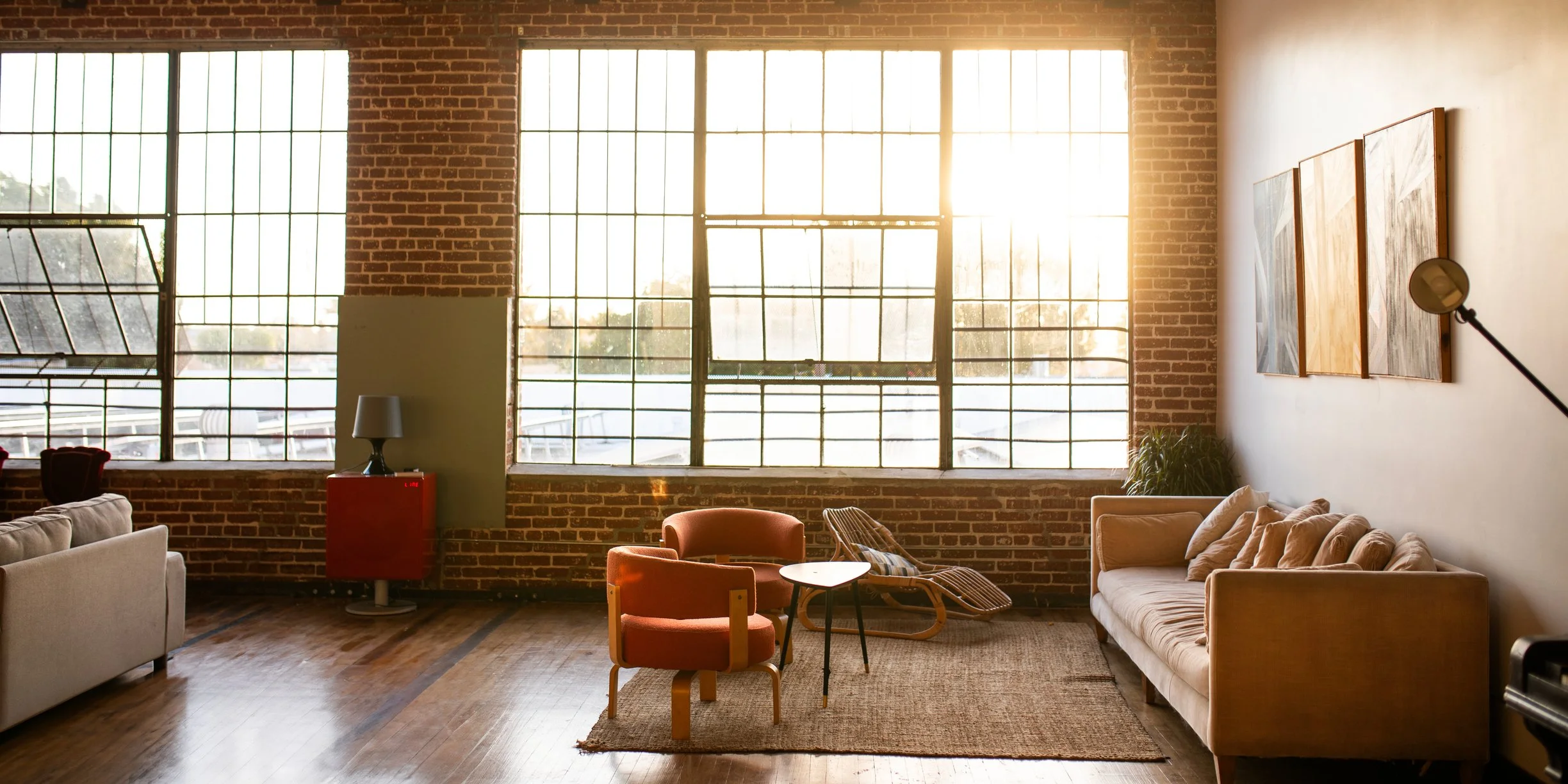 A spacious living room with large brick wall windows allowing sunlight to fill the room. The room features minimalist furniture including beige and orange armchairs, a beige couch with cushions, a small round table, and a red cabinet with a lamp. There are paintings on the right wall and a floor lamp near the artwork. The floor is wooden with a rug under the seating area.