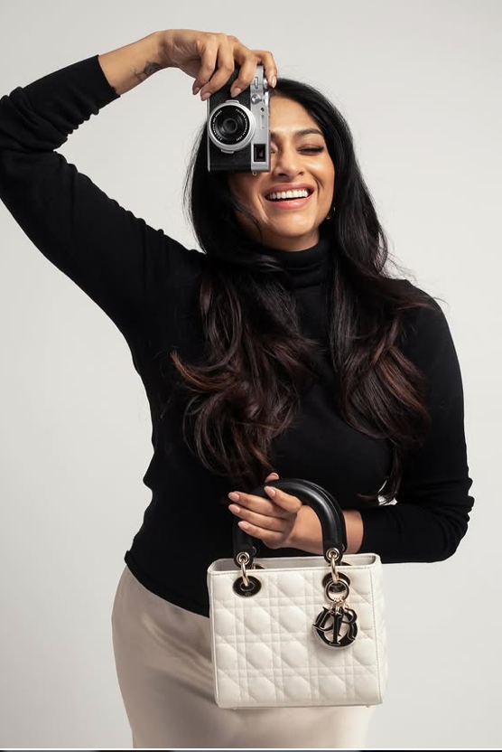 A woman with long wavy dark hair, smiling, holds a camera above her head with one hand and a white purse with gold hardware in the other hand, standing against a plain light gray background.