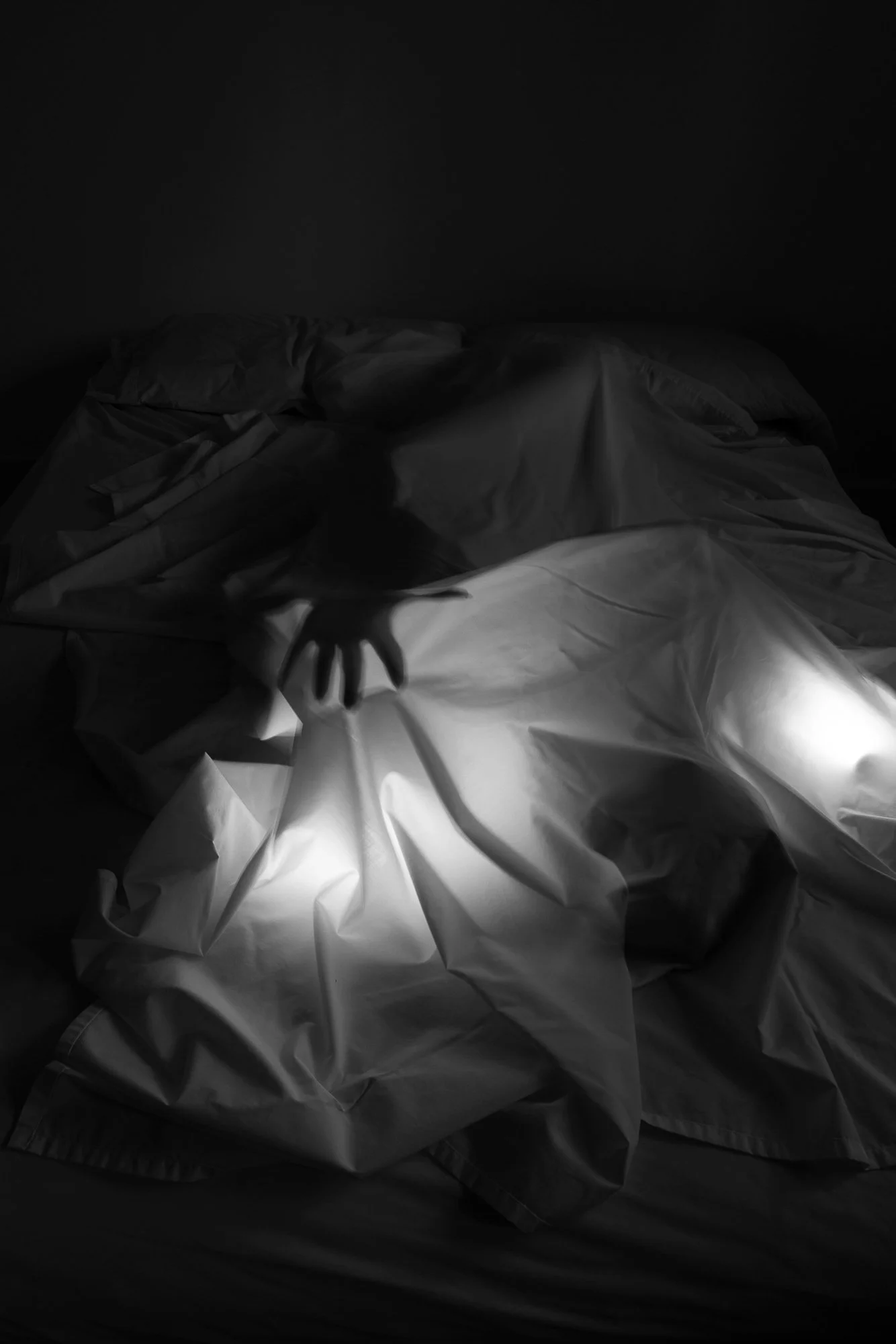 A darkened bedroom with a bed covered in crumpled white sheets and blankets. A small hand is reaching out from under the covers, illuminated by a faint light.