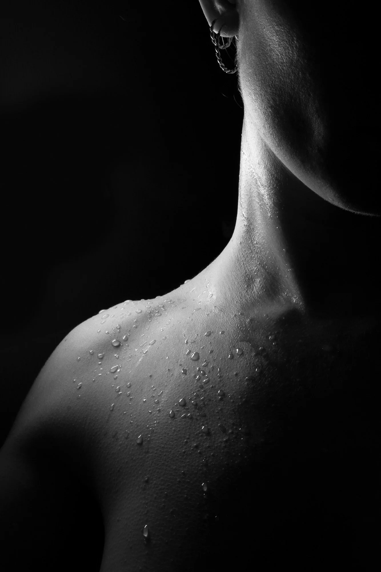 Close-up black and white photograph of a person's neck and shoulder with water droplets on the skin, partial view of face with lips slightly parted, dark background.