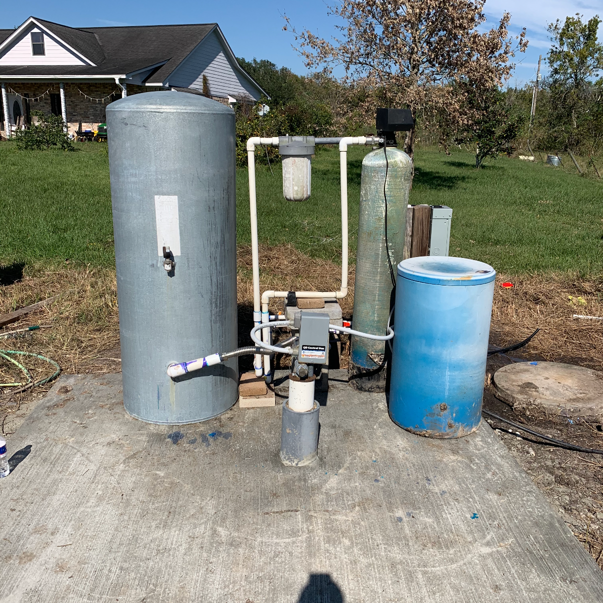 A backyard water treatment system setup with a large gray tank, a water filtration system, and a blue water storage barrel on a concrete slab, with a house and trees in the background.