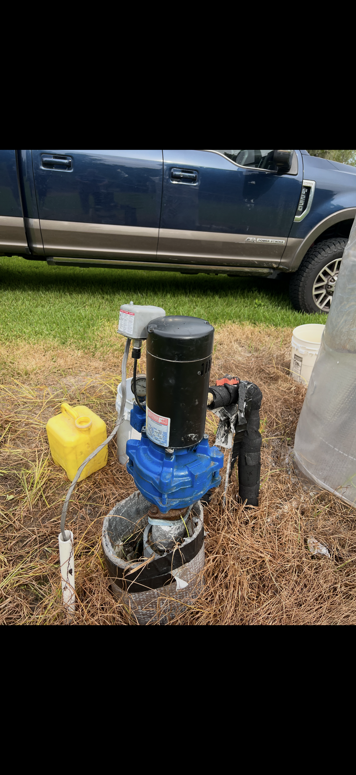 A water well pump with a blue motor, black pipe, and yellow fuel container on the ground in front, set in a grassy area with a dark blue pickup truck in the background.