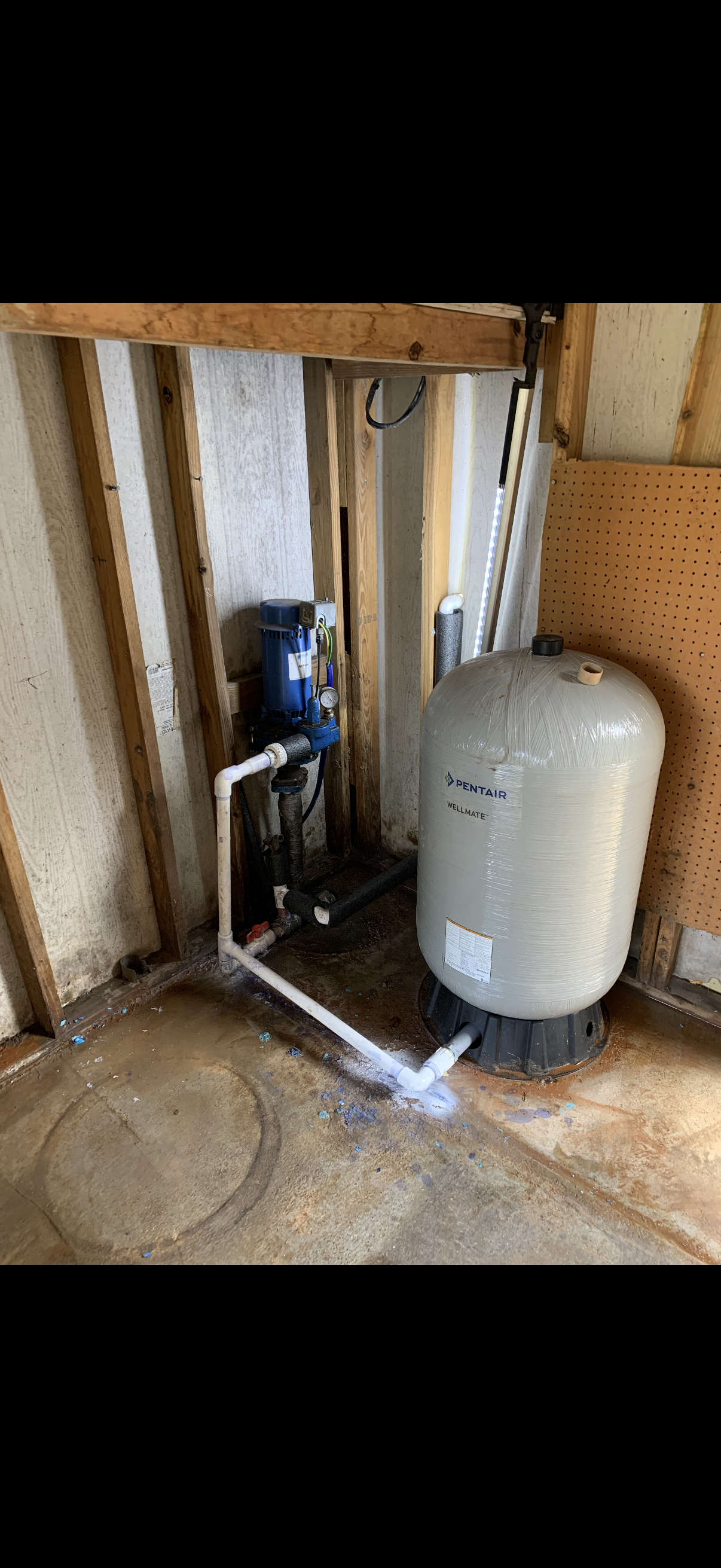 A water filtration and softening system installed in a basement with exposed wooden framing, a large white Wellmate water tank, and plumbing pipes.