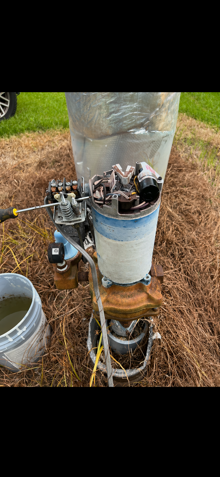 Close-up of an old, partially disassembled water pump with wiring and mechanical parts, surrounded by dry grass and a bucket of water nearby.