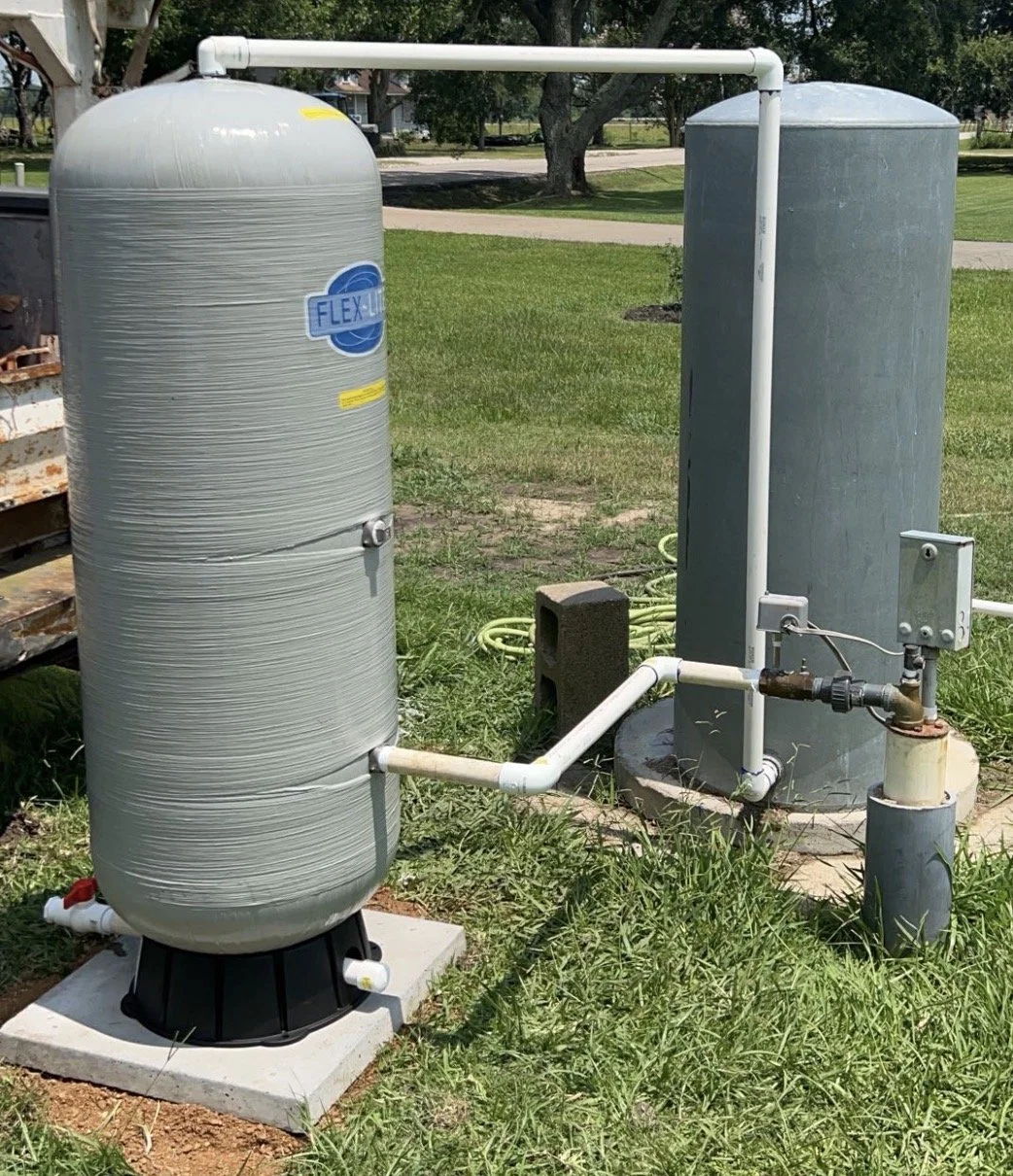 Outdoor water filtration system with large gray tanks connected by white pipes on grass, with trees and a sidewalk in the background.