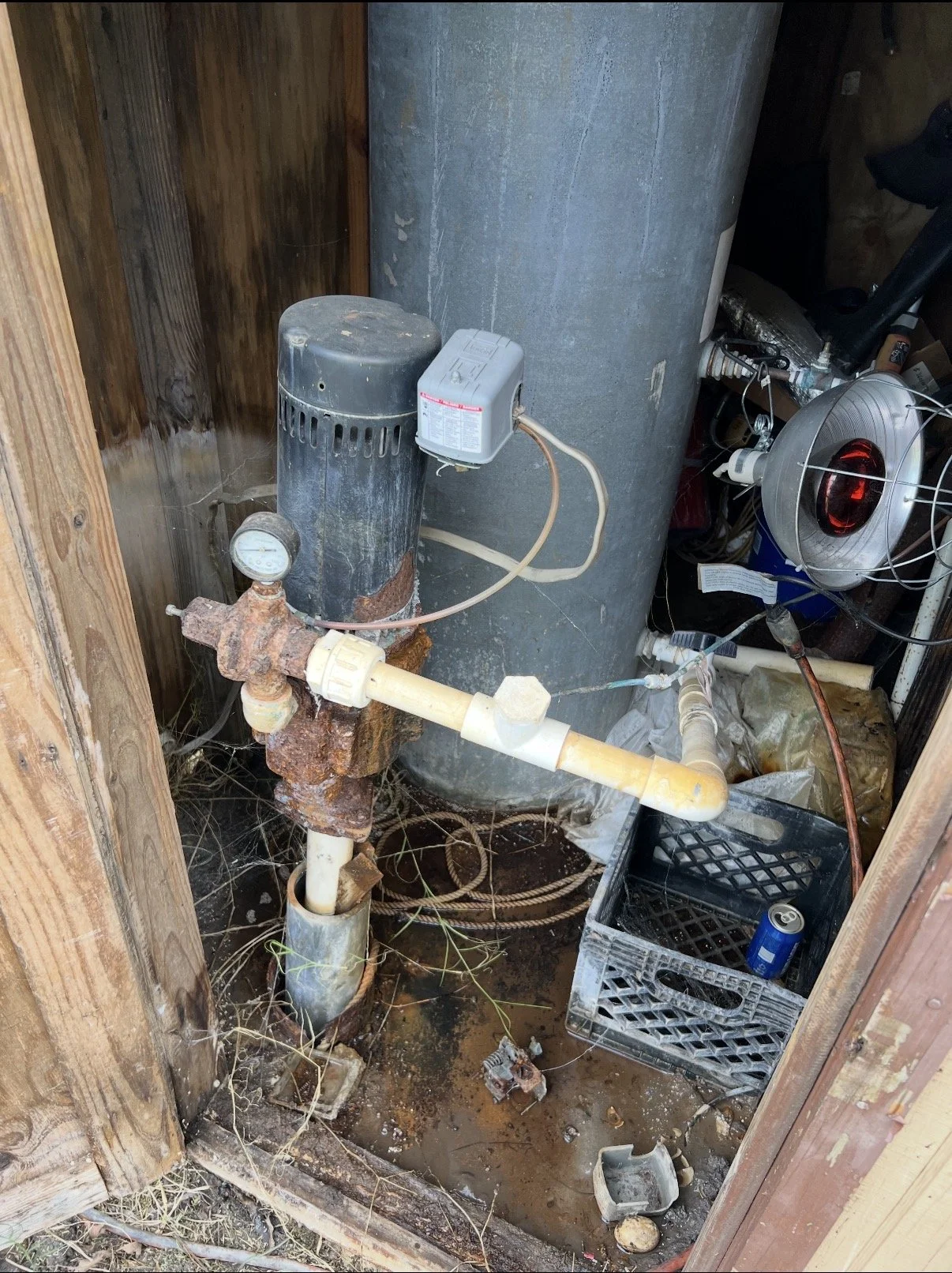A small, cluttered utility space with rusted plumbing, an aged pressure tank, and a rusty valve connected to pipes. The floor is dirty with debris, dirt, and cobwebs. There’s a black plastic crate with a can and some miscellaneous items. The space appears unkempt and cluttered.