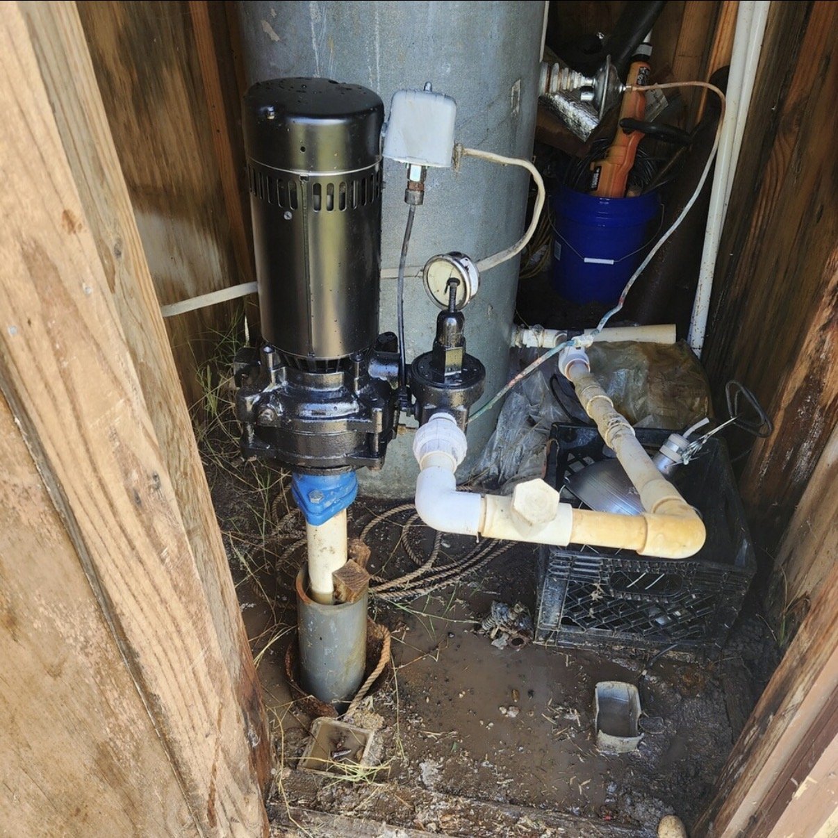 A sump pump system installed in a small pit, with pipes, wires, and a blue bucket containing tools nearby, inside a wooden enclosure.