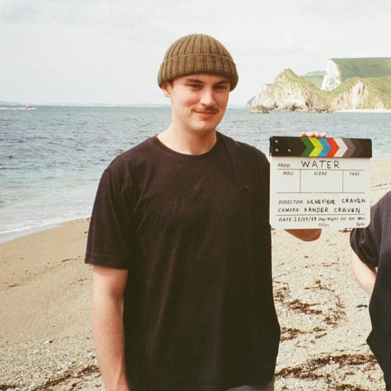 Young man in black t-shirt and brown beanie holding a film clapperboard on a beach with cliffs in the background.