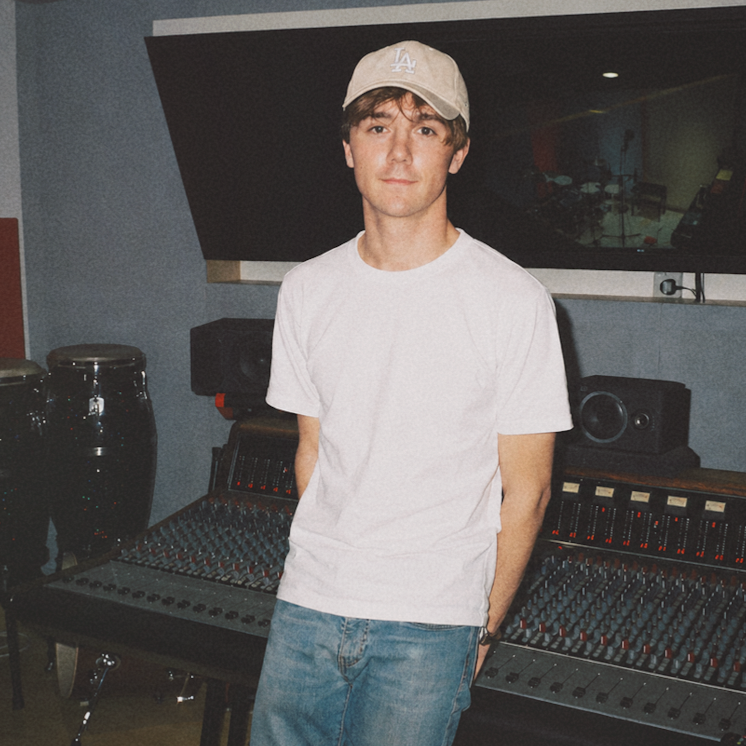 A young man in a light-colored baseball cap and white t-shirt standing in a recording studio, with audio equipment and mixing consoles around him.