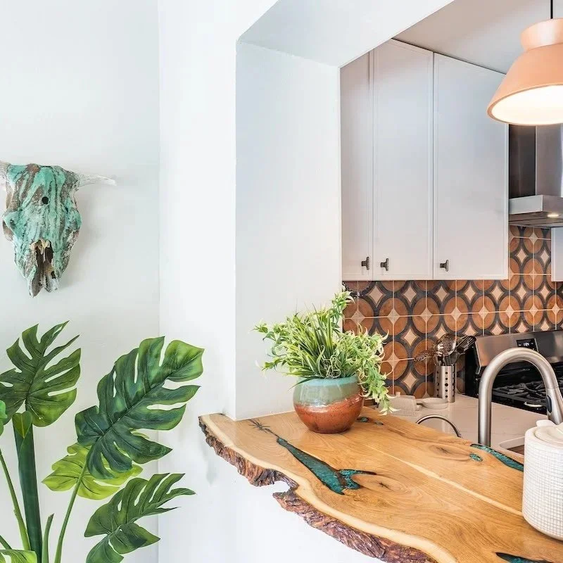 Interior view of a modern kitchen with a wooden countertop, white cabinets, decorative tile backsplash, houseplants, and a hanging light fixture.