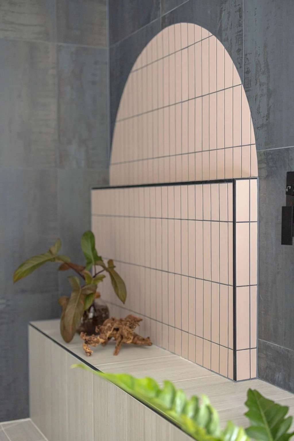 Arched tile installation with multiple colors and textures on primary bathroom shower wall with ledge and plants.