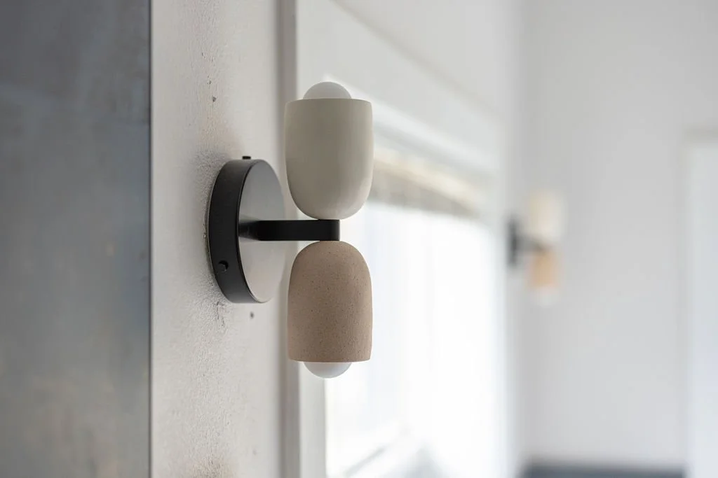 Modern wall-mounted light fixture with two geometric shades, one white and one beige, attached to a black circular base, in a bright room with natural light.