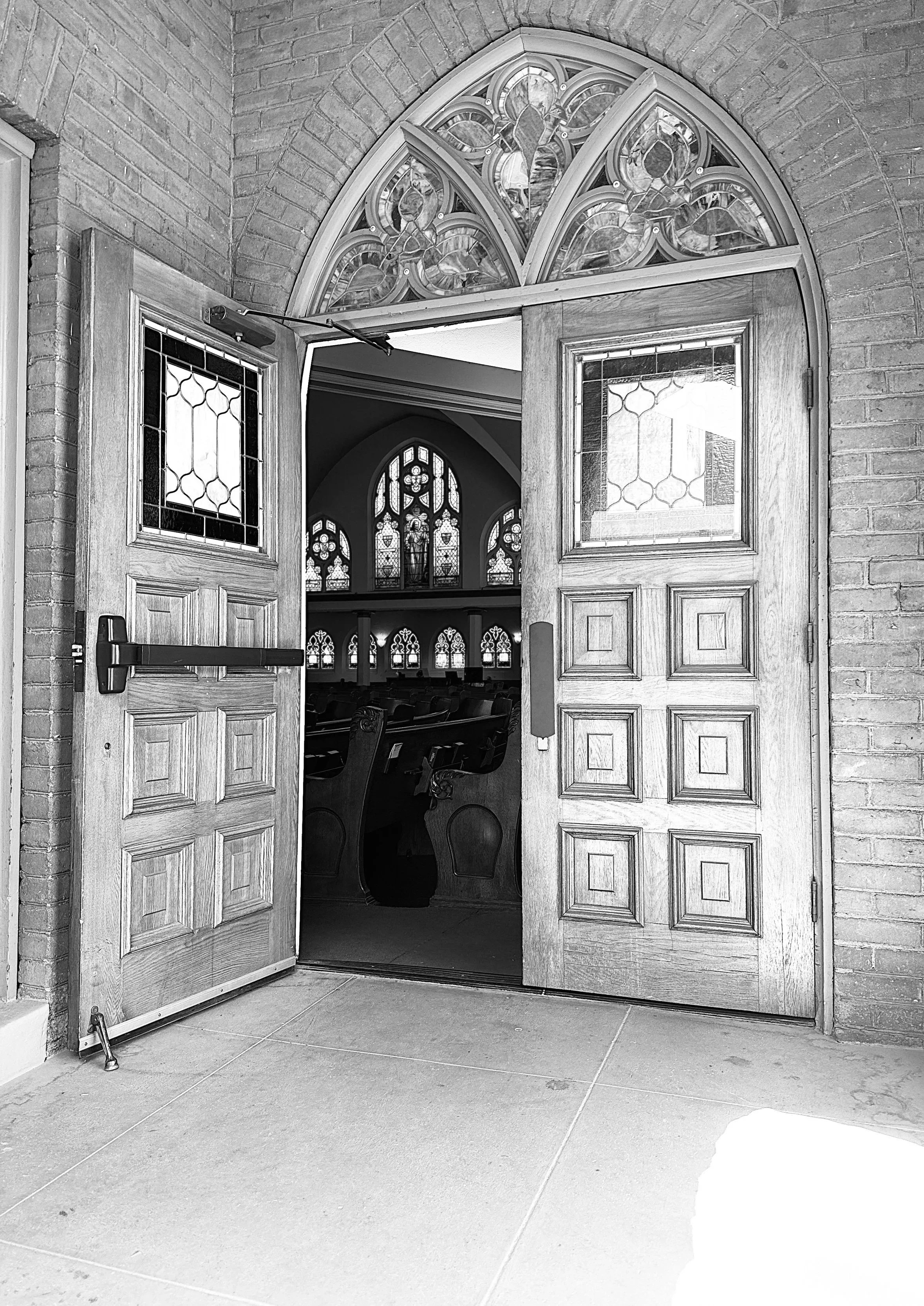 Wooden church doors open inward, revealing stained glass windows and pews inside. The doors have decorative stained glass panels, and above the doorway is a gothic-style stained glass window set in a brick wall.