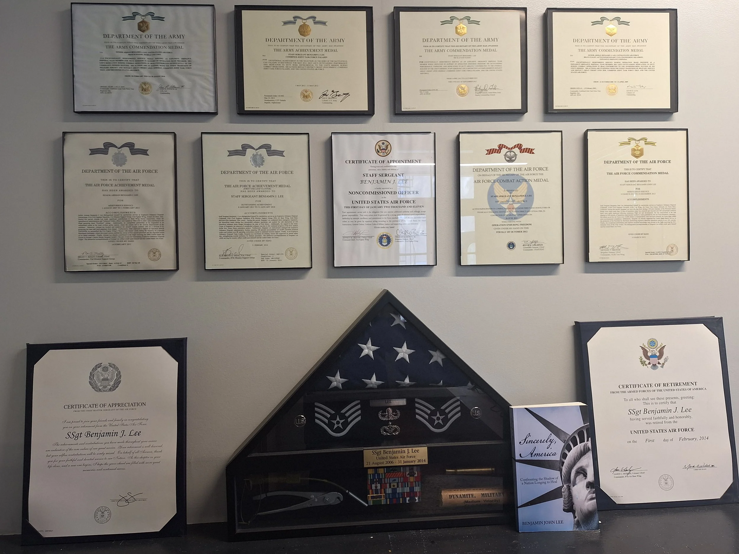 Display of multiple military awards, medals, certificates, and commendation plaques on a wall, including medals from the Department of the Army and the United States Air Force, along with framed recognition documents and a folded American flag in a box.