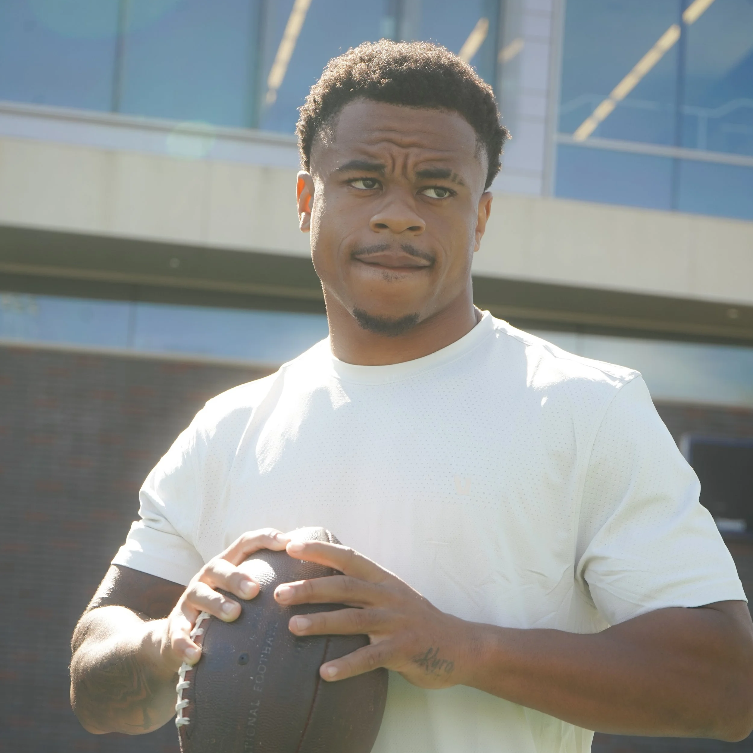 A young man with curly hair holding a football, outdoors on a sunny day with a modern glass building in the background.