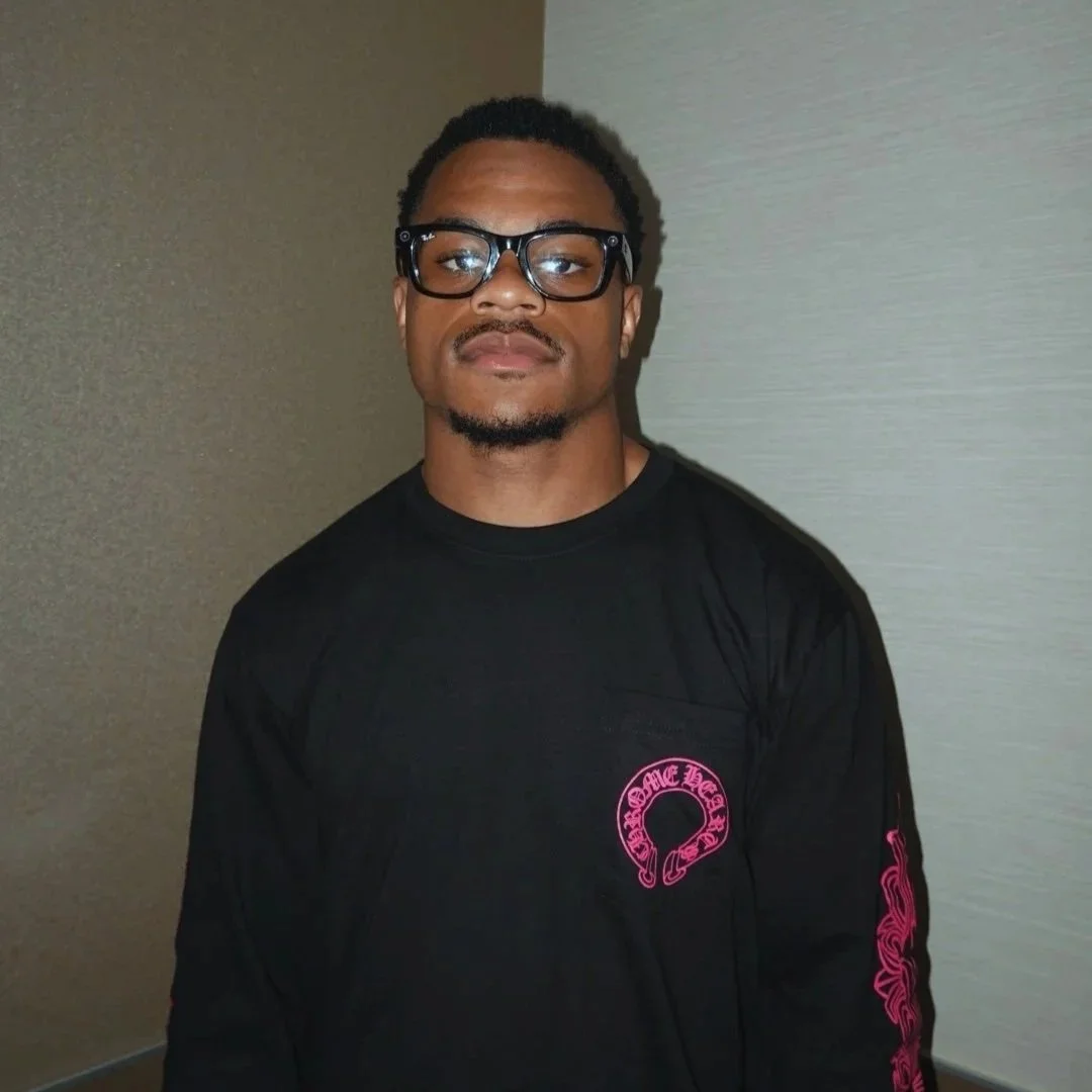 A young man with glasses, short curly hair, and a goatee wearing a black shirt with pink designs standing against a beige wall.