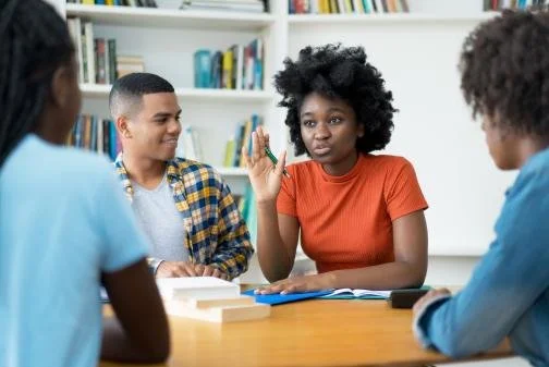 Four young students sitting at a table in a library or classroom, engaged in a discussion. One girl with curly hair and an orange shirt is speaking and raising her hand, while the three boys listen.