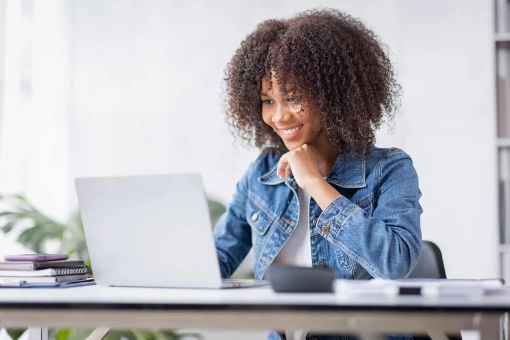 A woman with curly hair working on a laptop at a desk in a bright room.