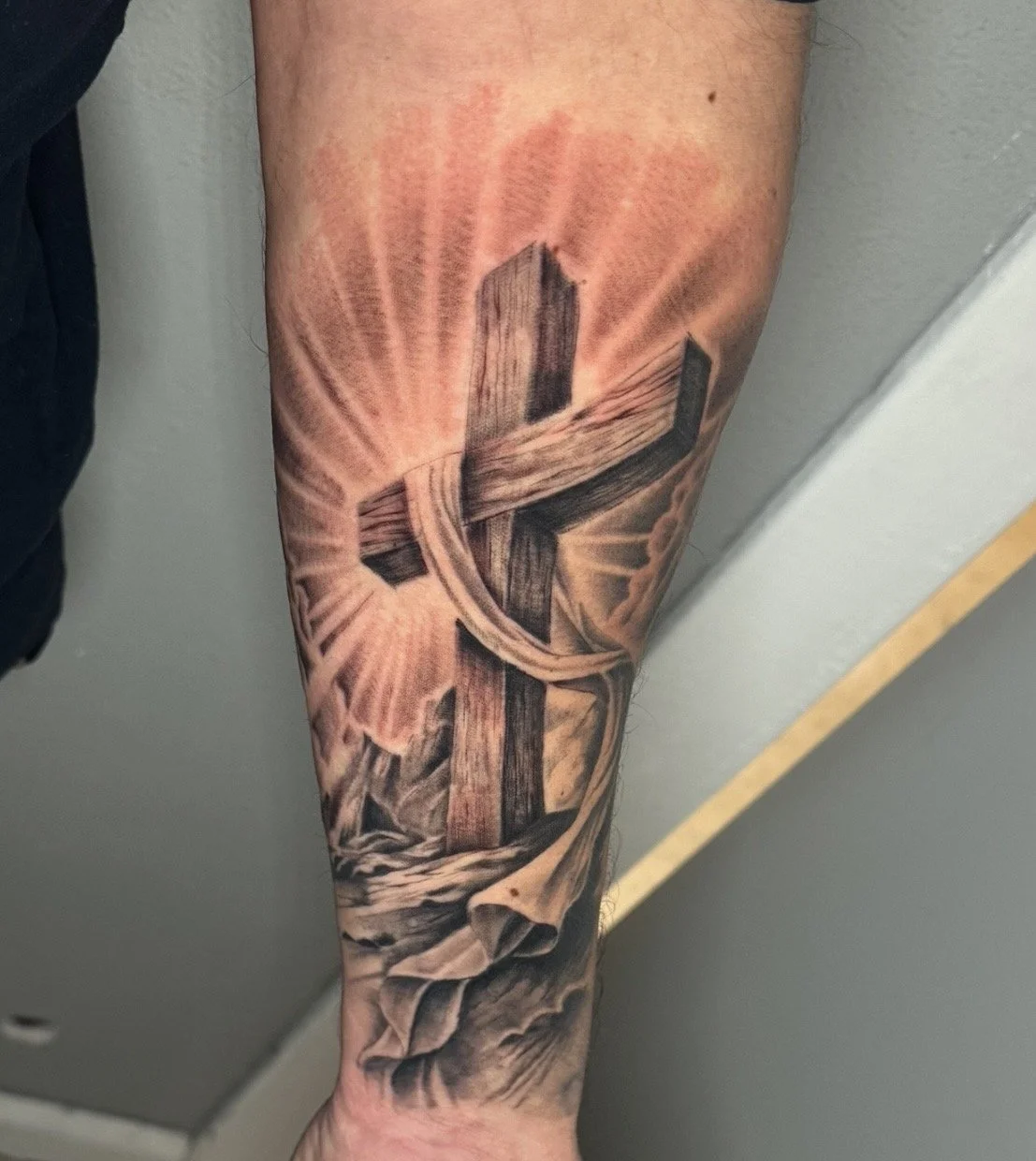 A realistic black and gray tattoo of a wooden cross with a cloth draped over it. The background features radiant light rays and clouds.