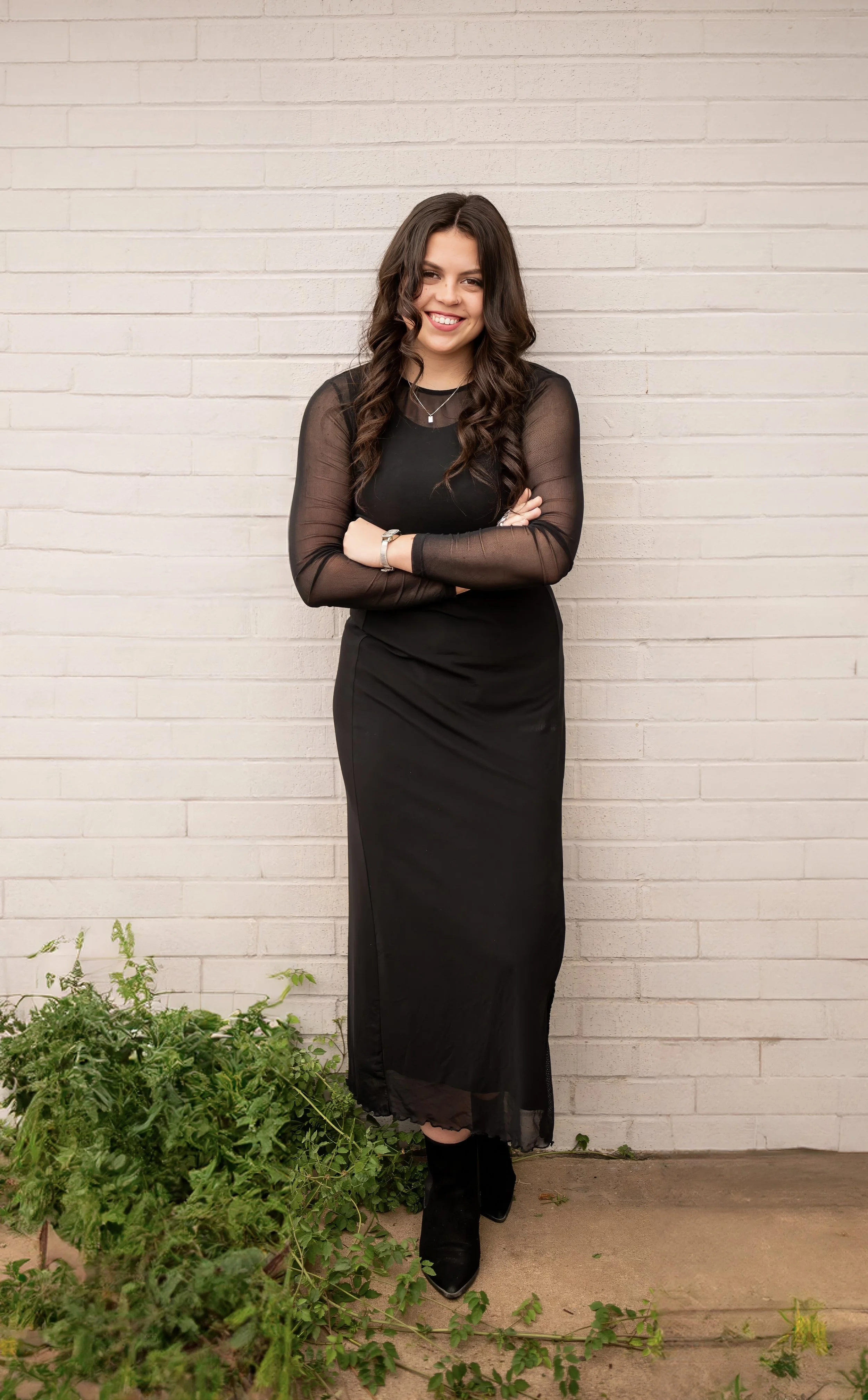 A woman with long dark wavy hair, wearing a black dress with sheer long sleeves and black boots, stands against a white brick wall with her arms crossed and smiling. There are green plants at her feet.