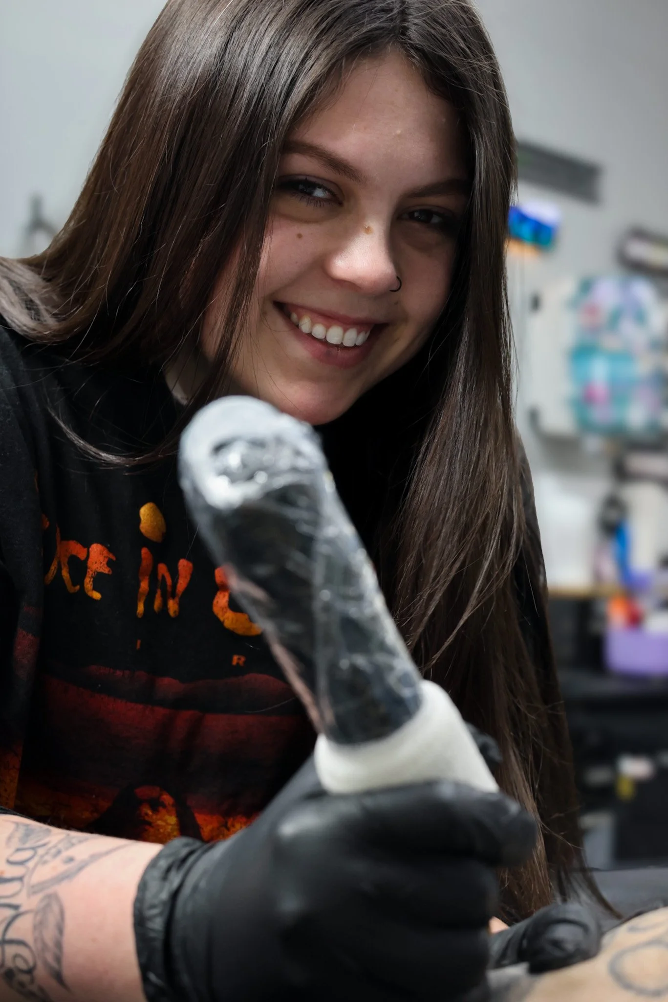 A young woman with long brown hair, nose piercing, and tattooed arm, smiling while tattooing on someone's arm in a tattoo studio.