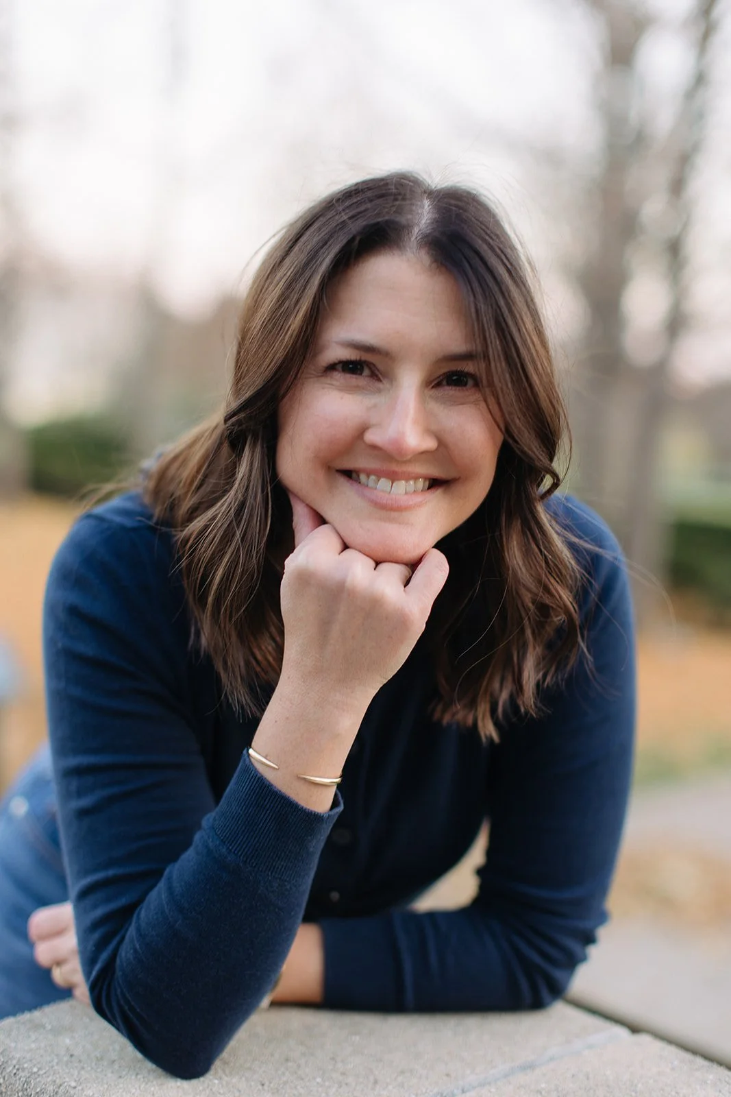 A woman with brown hair smiling, leaning on her hand outdoors during fall, wearing a navy blue top, with trees and blurred background.