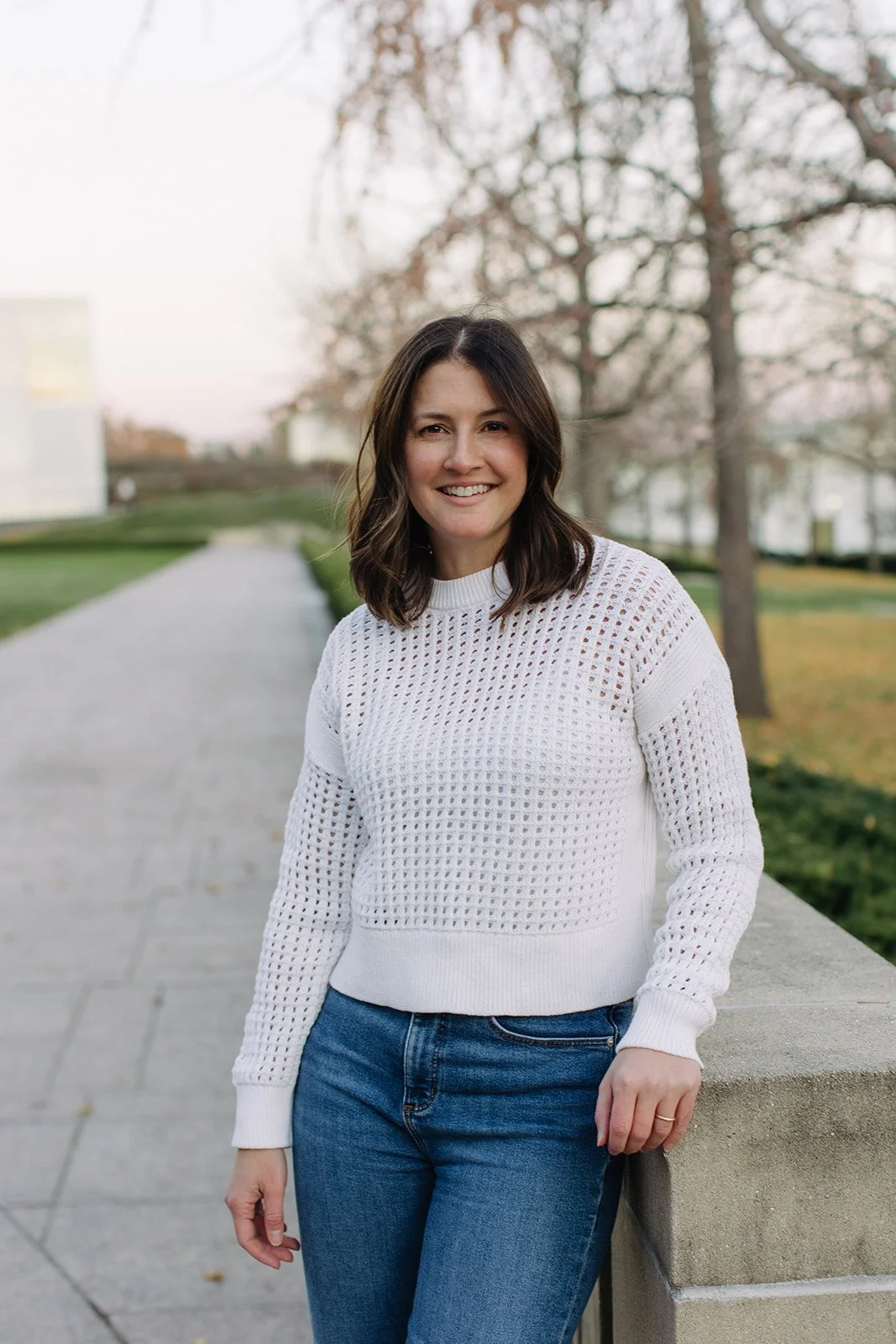 A woman with shoulder-length dark hair smiling at the camera outdoors, wearing a white open-knit sweater and blue jeans, standing next to a stone railing with trees and buildings in the background during daylight.