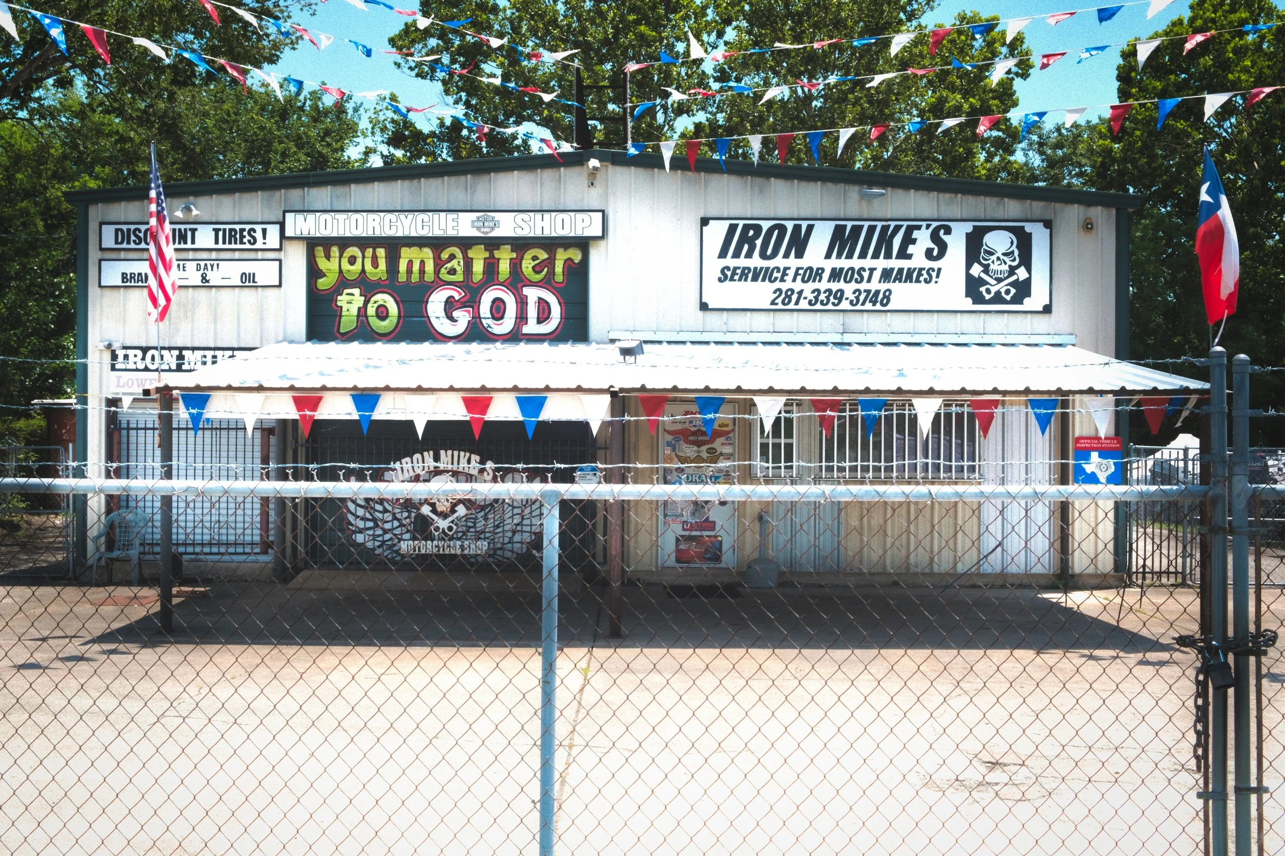 A motorcycle shop with signs and banners, including American flags and red, white, and blue pennants, behind a chain-link fence, with trees and blue sky in the background.