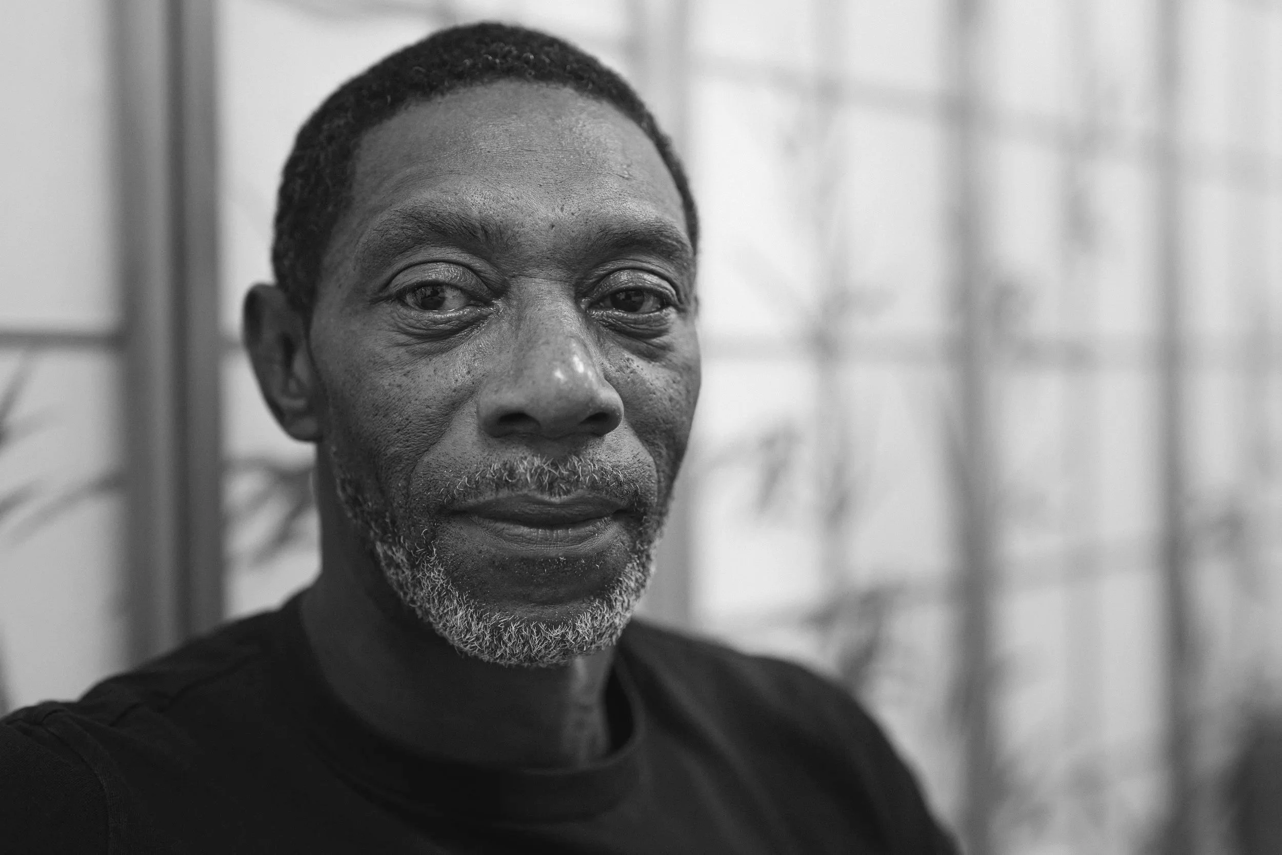 A close-up black-and-white portrait of an older African American man with short hair and a grayed beard, wearing a dark shirt, looking directly at the camera with a neutral expression.