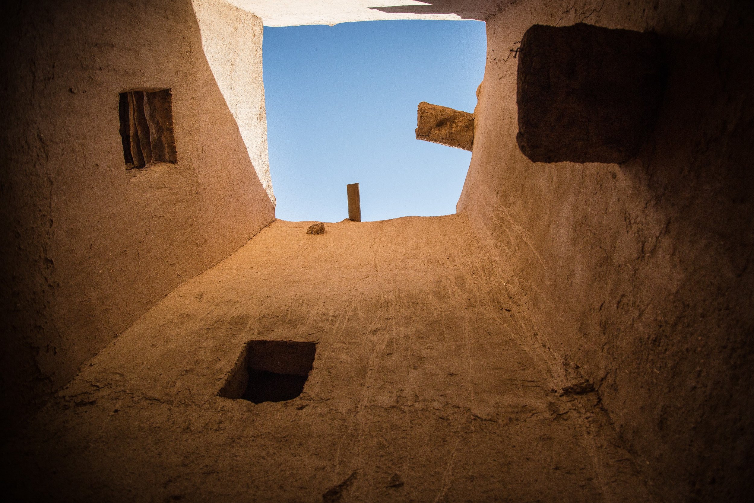Looking up from inside a ruined structure with rough beige walls, through an opening at the top revealing a clear blue sky, with stones and debris at the bottom.