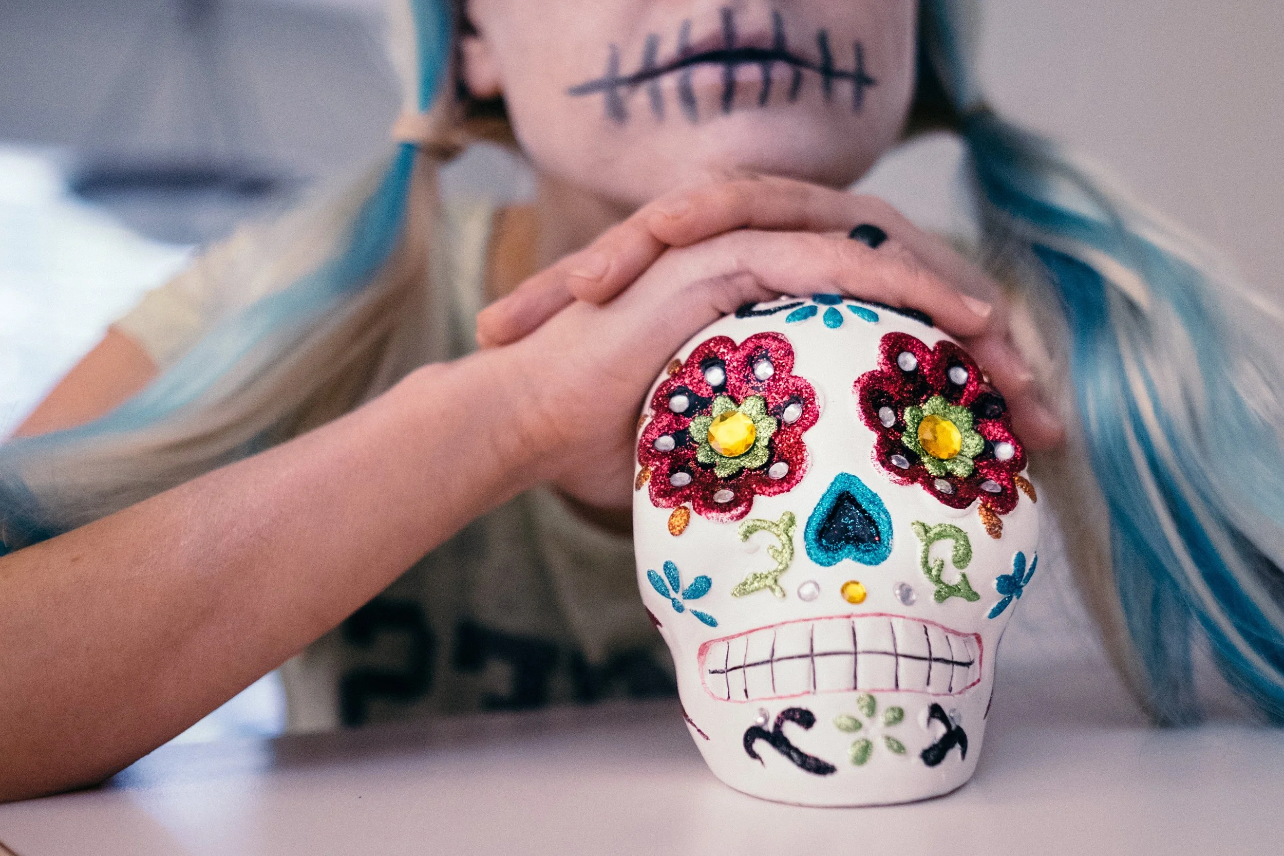 Hand-painted decorative skull with colorful floral and geometric designs, inspired by Día de los Muertos, held by a person with facial makeup resembling a sugar skull.