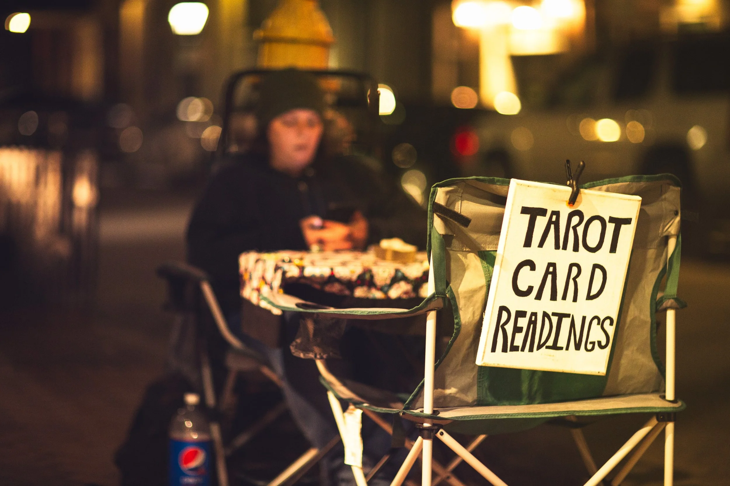 A person sitting at a table at night with a sign that says 'Tarot Card Readings' in front of them. The scene is blurred with warm lights in the background.