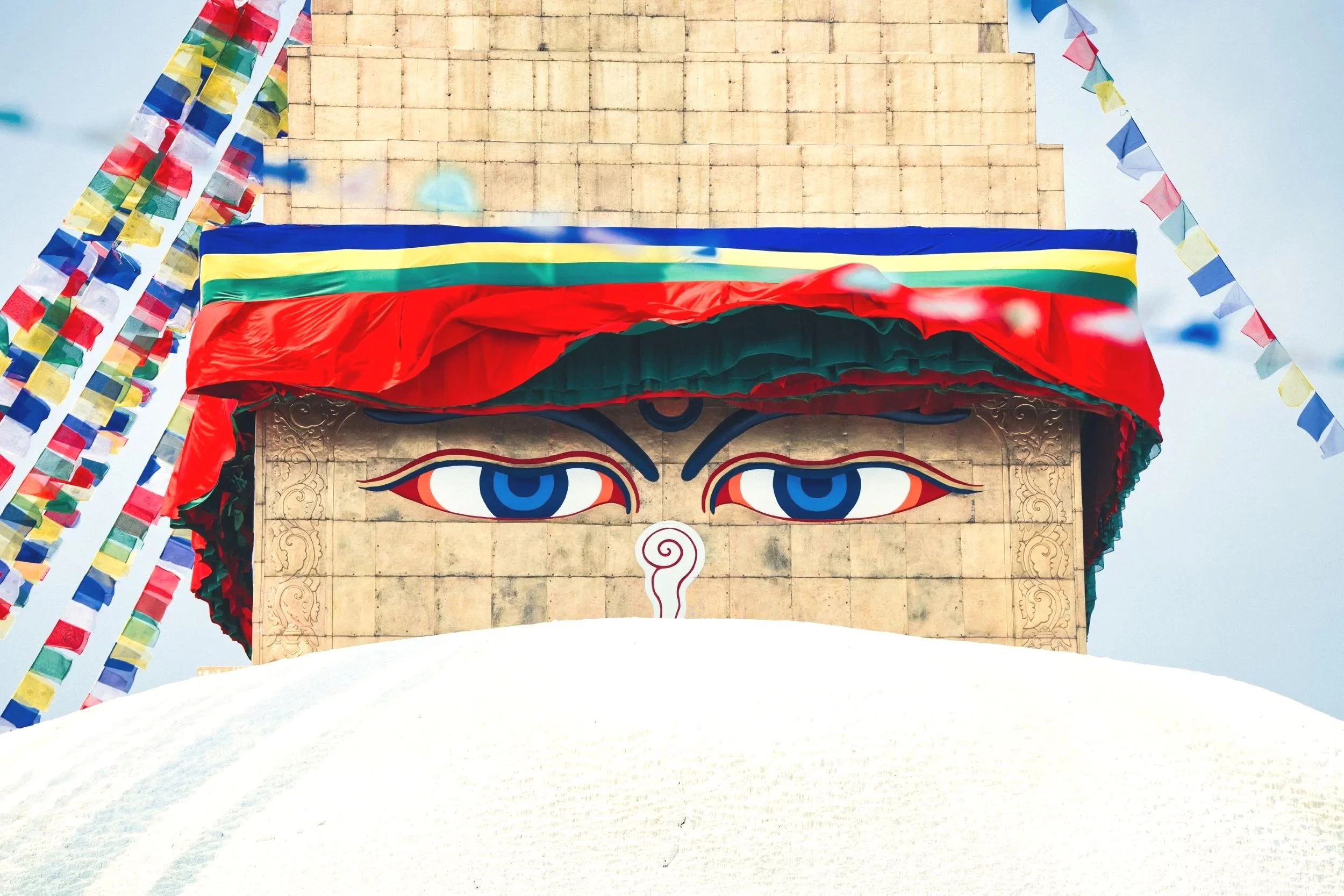 Close-up of a colorful, decorative face featuring large blue eyes, red, green, and yellow accents, and traditional design elements, surrounded by vibrant prayer flags in red, blue, green, yellow, and white fluttering against a light sky. The background includes a beige stone wall with horizontal lines.