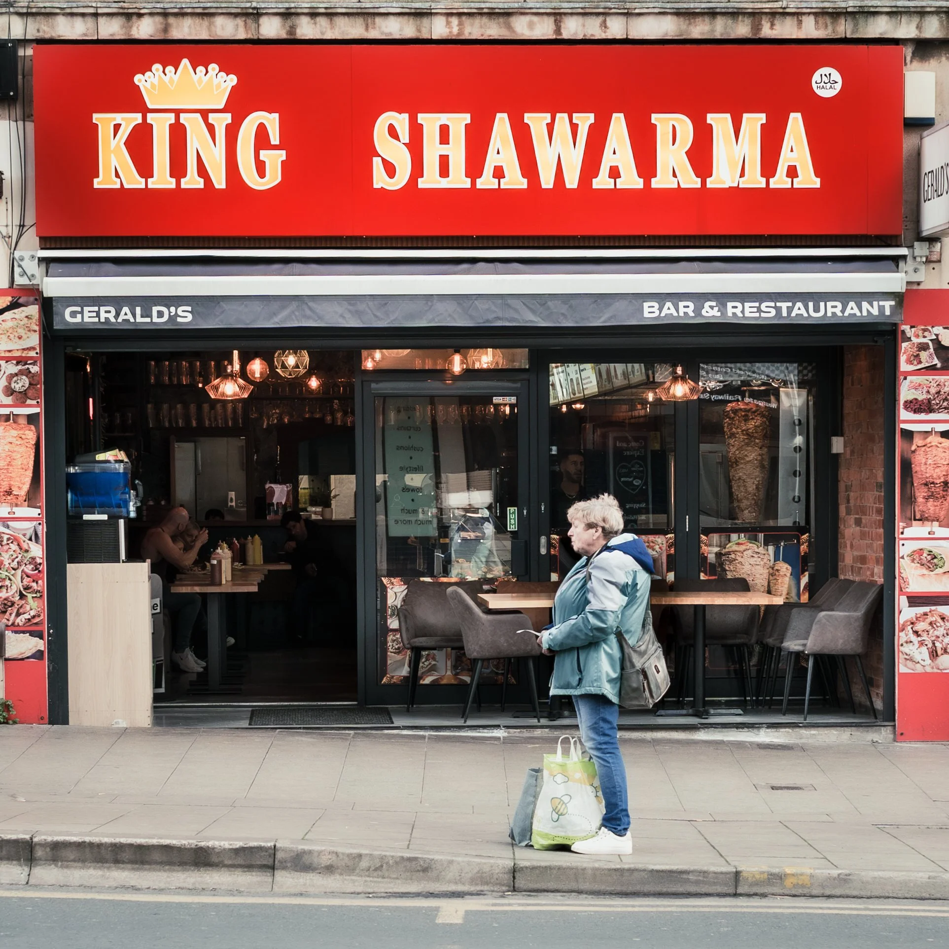 Front view of King Shawarma restaurant with a large red sign and logo above the entrance, two people sitting inside, and a man standing outside with a shopping bag.
