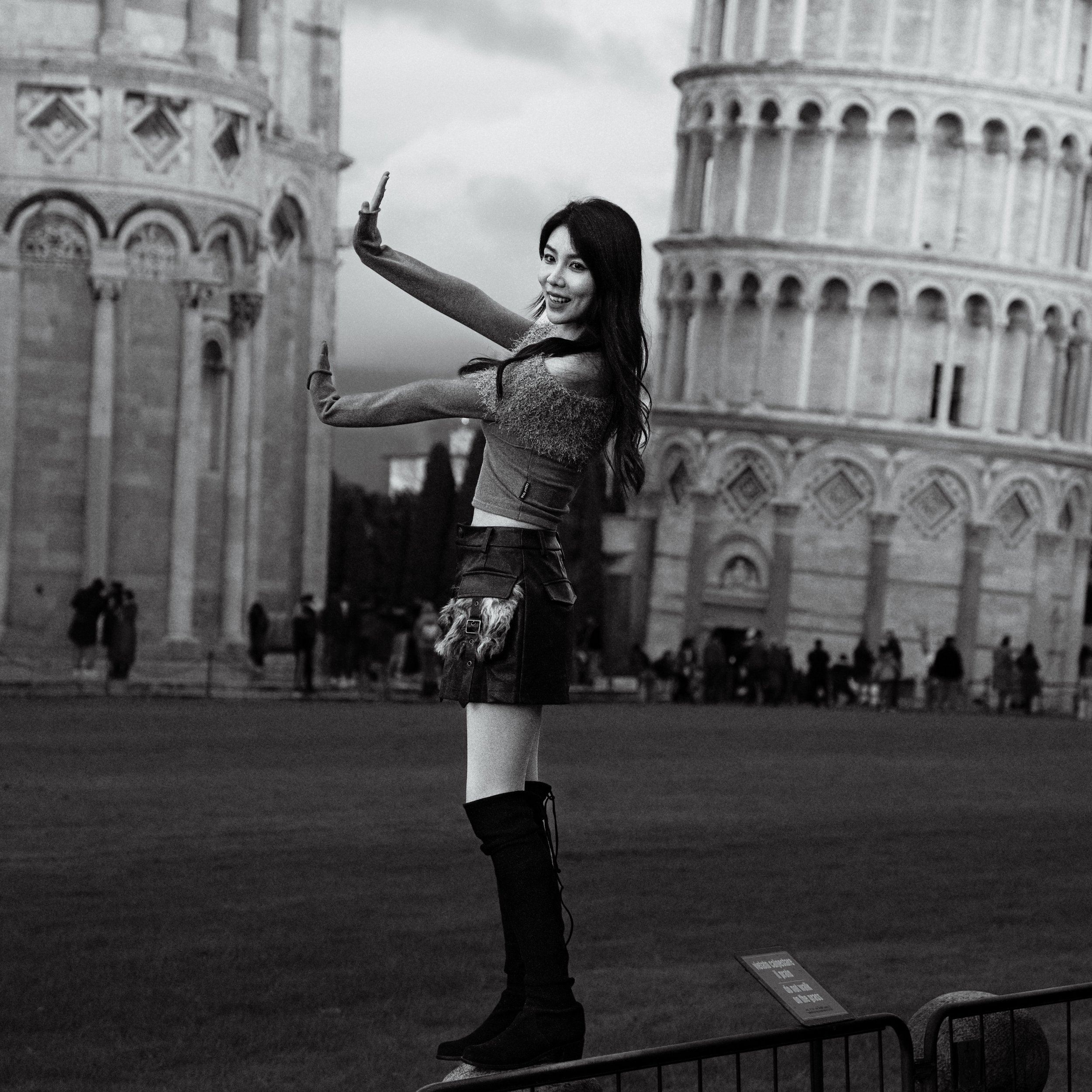 A woman standing on a pedestal in front of the Leaning Tower of Pisa, posing with her arm raised as if holding the tower, in black and white.