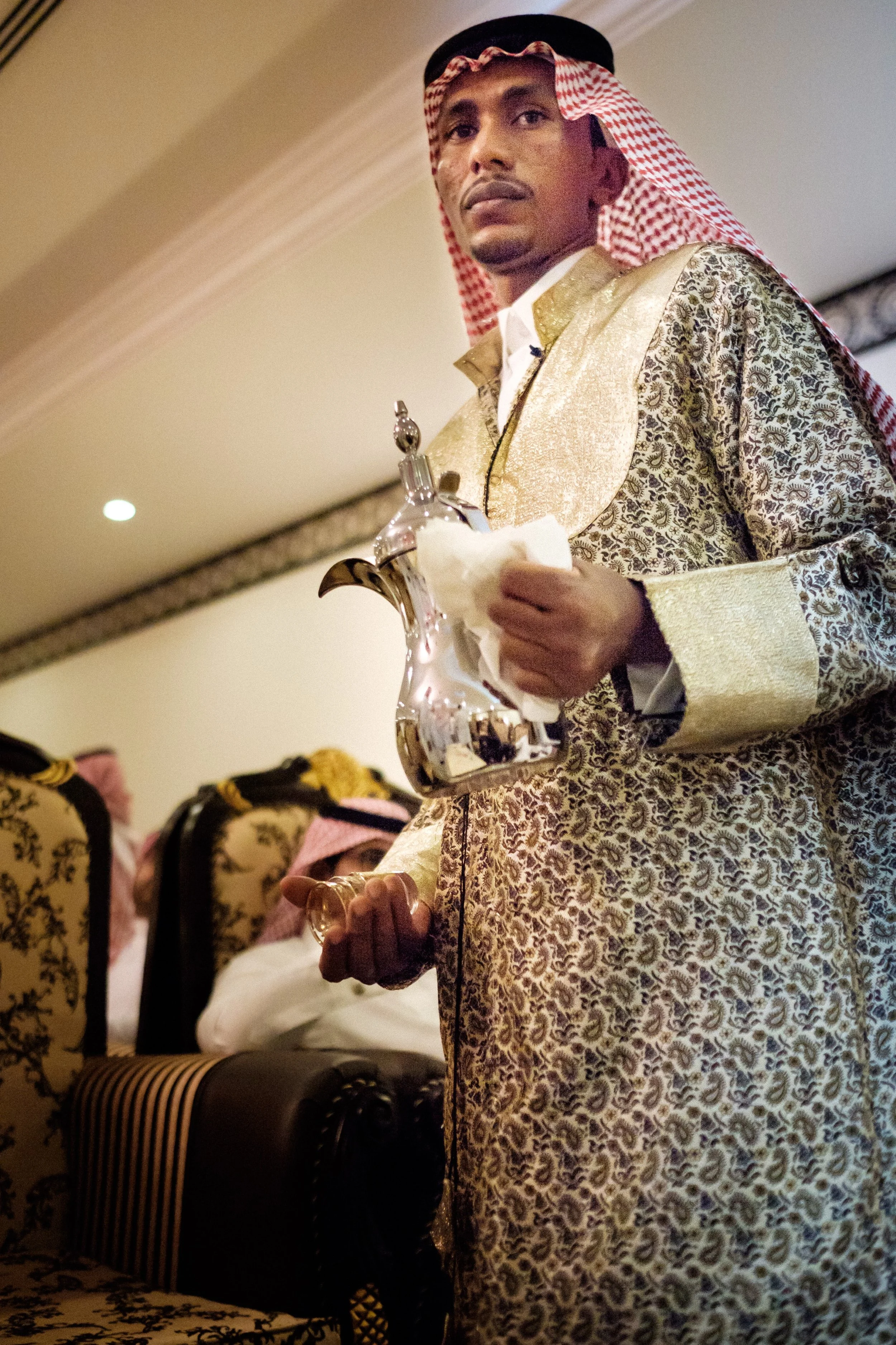 Man wearing traditional Middle Eastern attire, including a gold-patterned thobe and a red and white checkered keffiyeh, holding a trophy or decorative object.