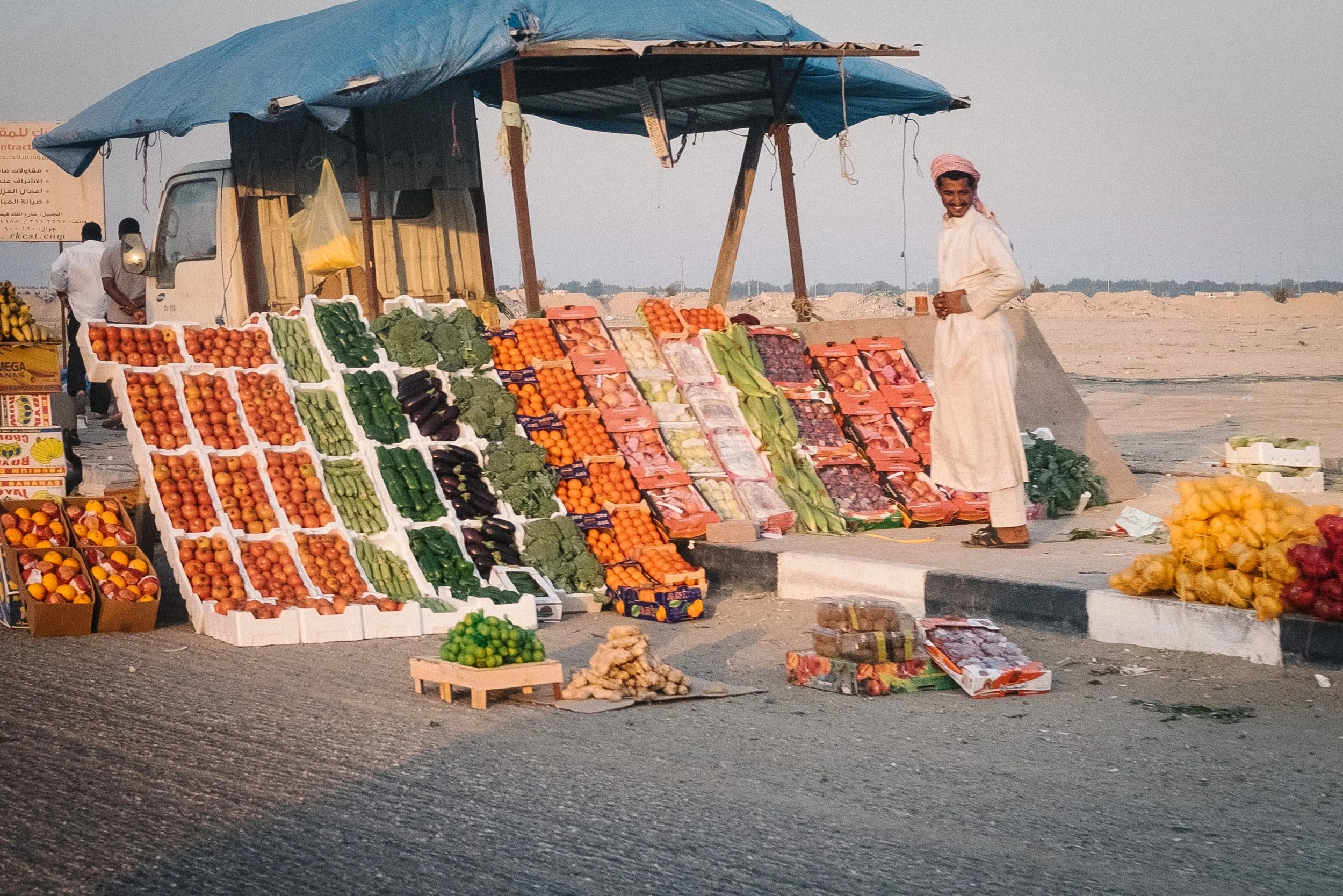 A street market stall with various fresh fruits and vegetables, including tomatoes, cucumbers, broccoli, oranges, and eggplants, under a blue tarp. A man in traditional white attire and a pink headscarf stands to the right of the stall, smiling, with