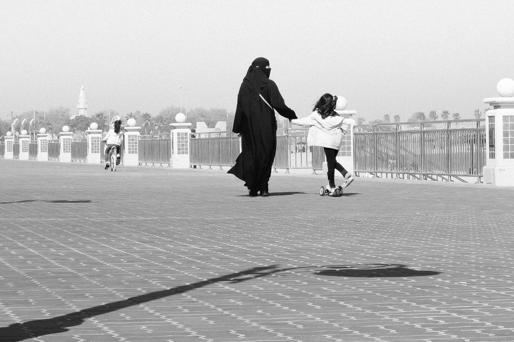 A woman dressed in traditional Islamic attire holding hands with a young girl as they walk on a paved area. In the background, a girl is riding a bicycle near a fenced area with decorative posts and spherical ornaments, under a clear sky with some trees and distant structures.