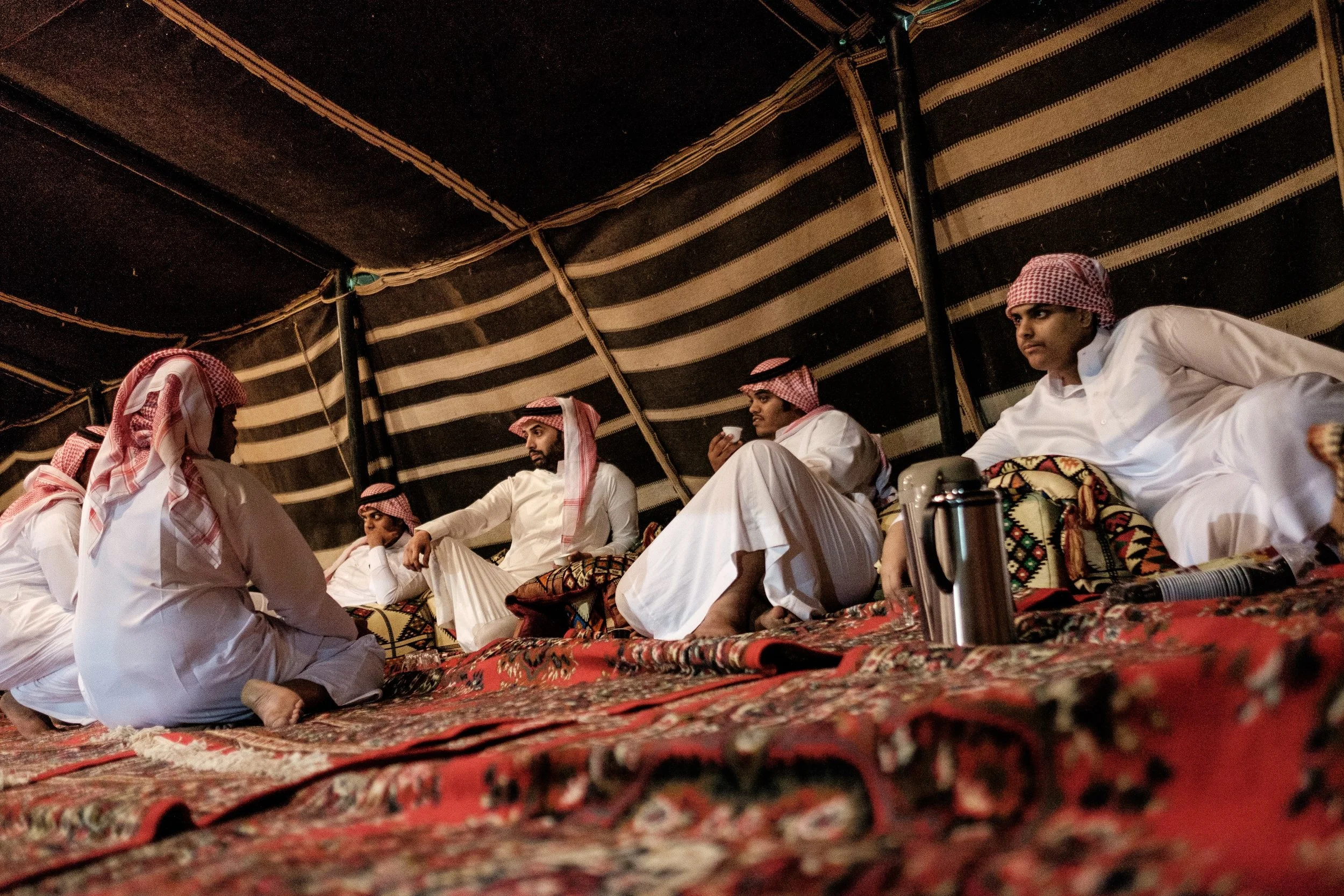 Several men in traditional Middle Eastern attire sitting cross-legged on a carpet inside a tent, with some holding cups and a thermos placed in front of them.