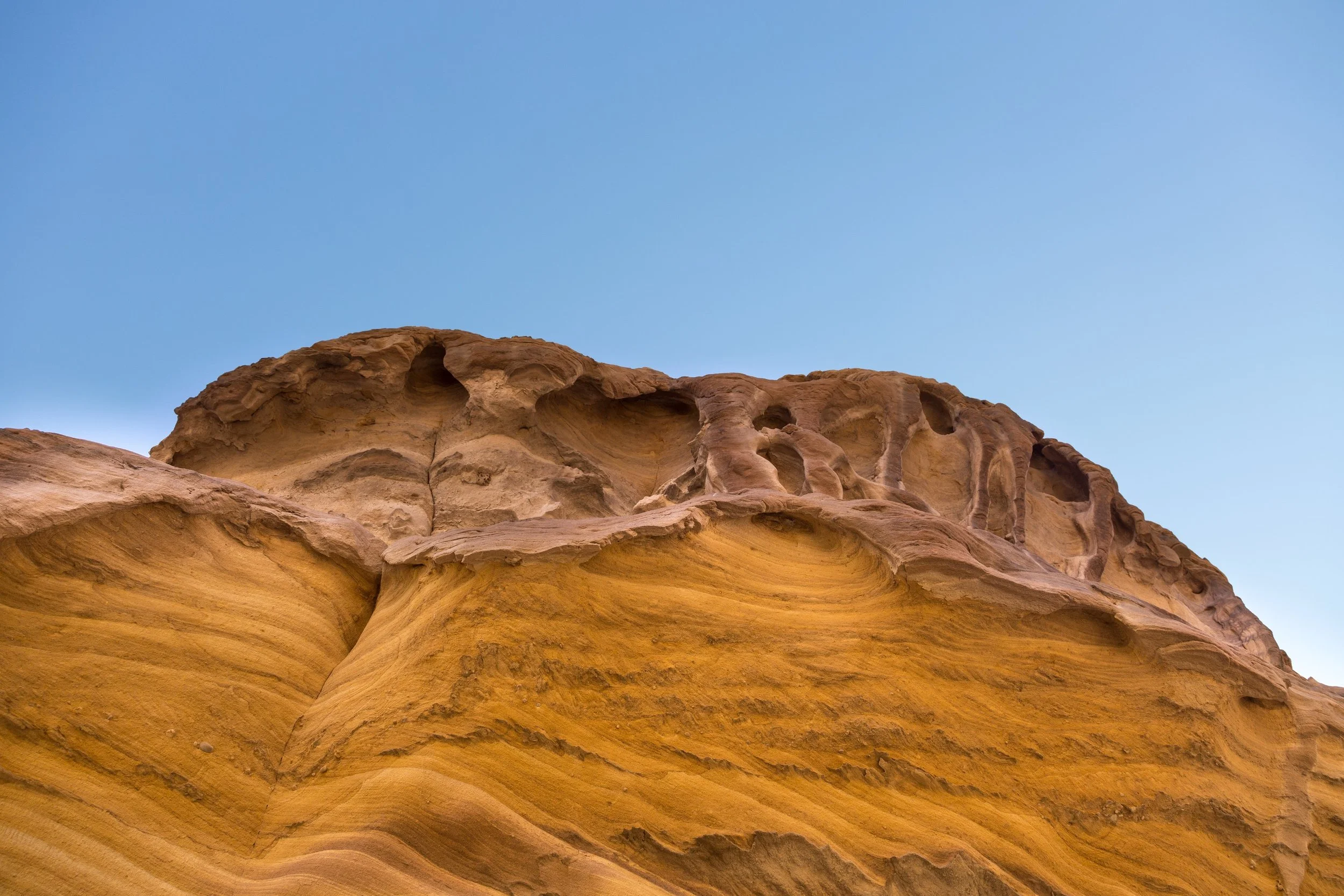 Close-up of a rugged, layered rock formation with warm orange and yellow tones, set against a clear blue sky.