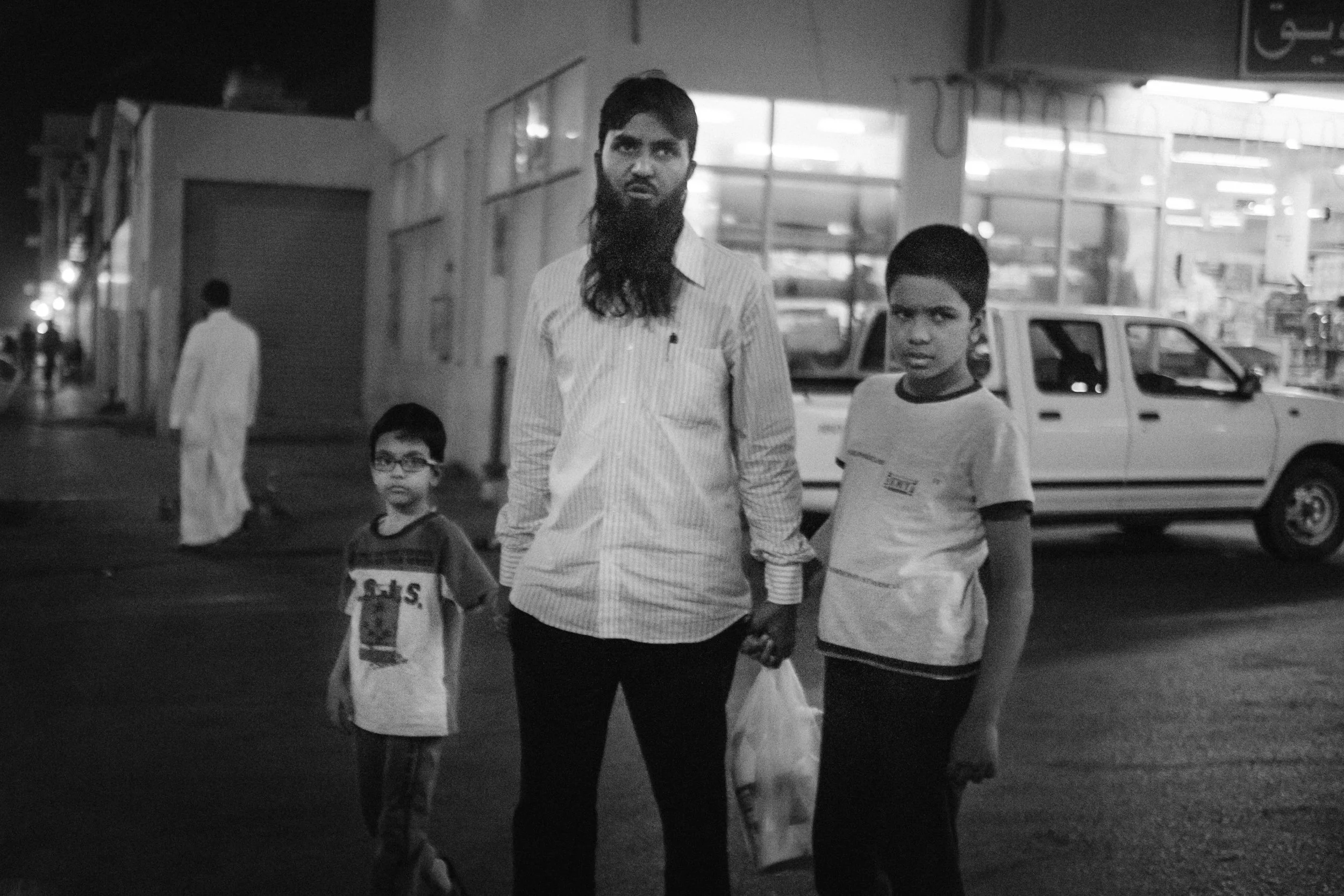 Black and white photo of two men and two children walking on a street at night, with cars and illuminated buildings in the background.