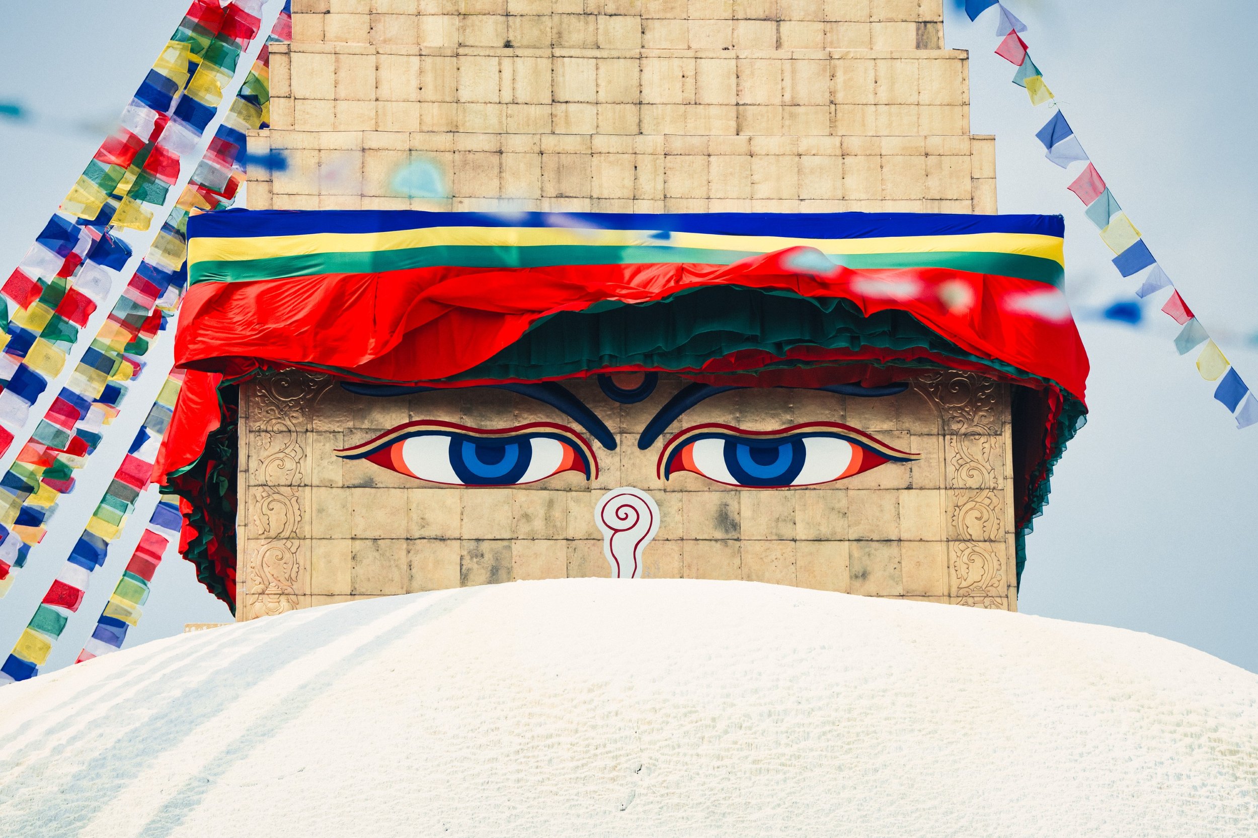 Close-up of a Buddhist prayer wheel with colorful 3D eyes and decorative fabric, surrounded by prayer flags, against a cloudy sky, on snow-covered ground.