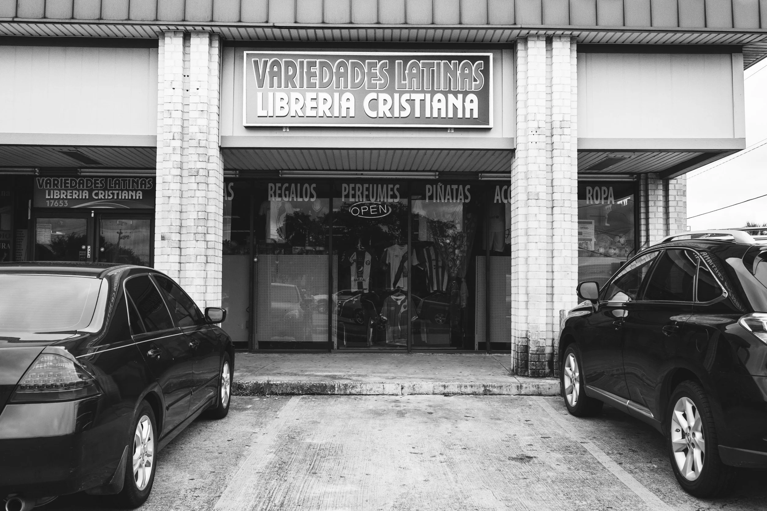 Black and white photo of a storefront with a sign that reads 'Variedades Latinas Libreria Cristiana.' There are cars parked in front of the store and the image includes the entrance and large windows with displays inside.